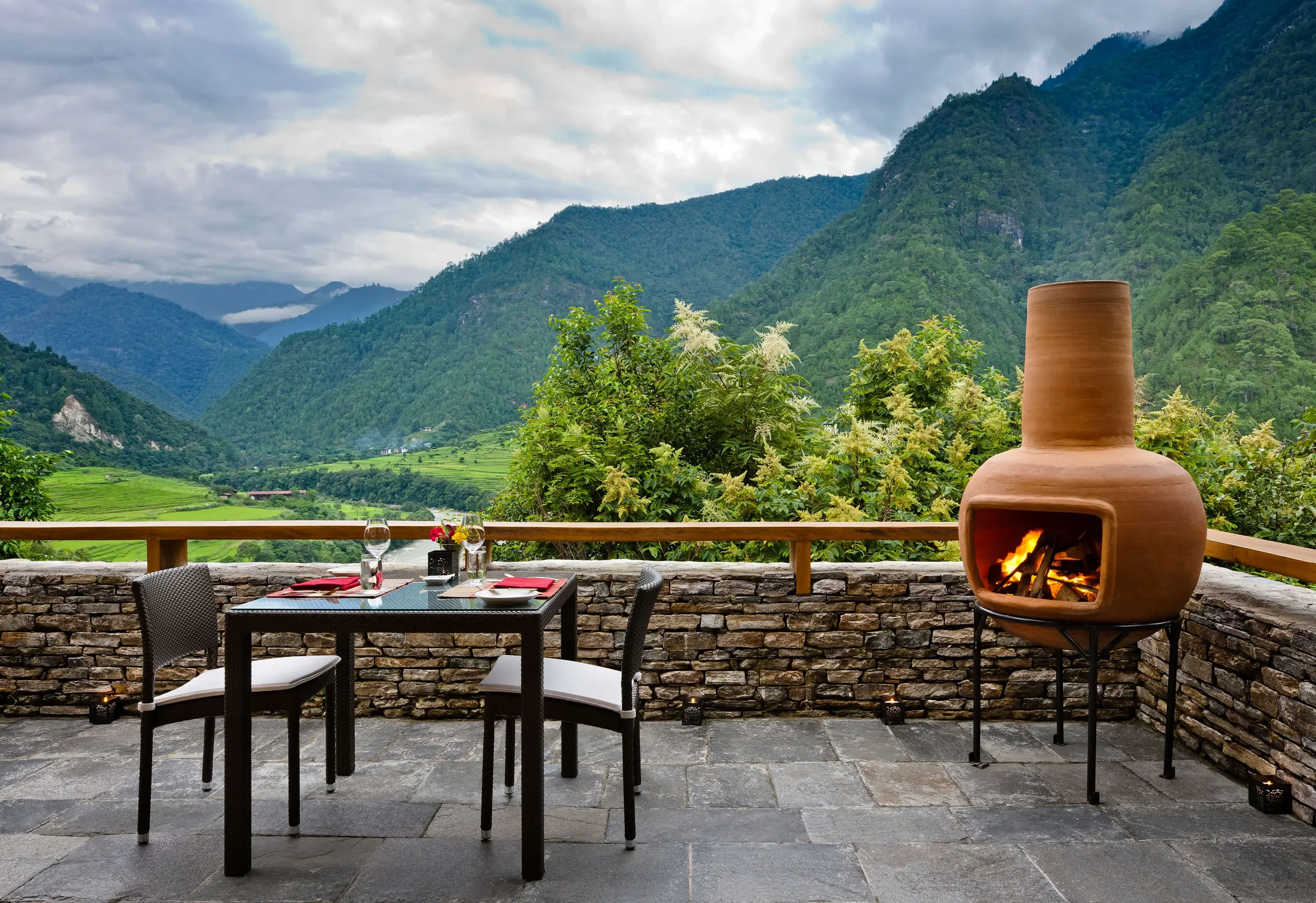 A scenic outdoor dining area with a mountain view, a fireplace, and a table set with glassware and red napkins.