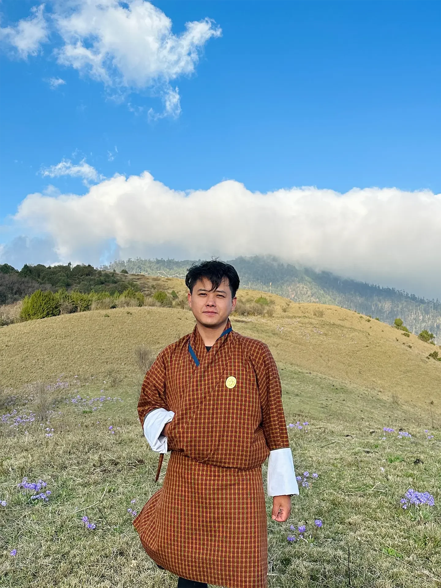 A young man in traditional clothing standing in a grassy field with purple flowers, mountains in the background, and a blue sky with clouds.