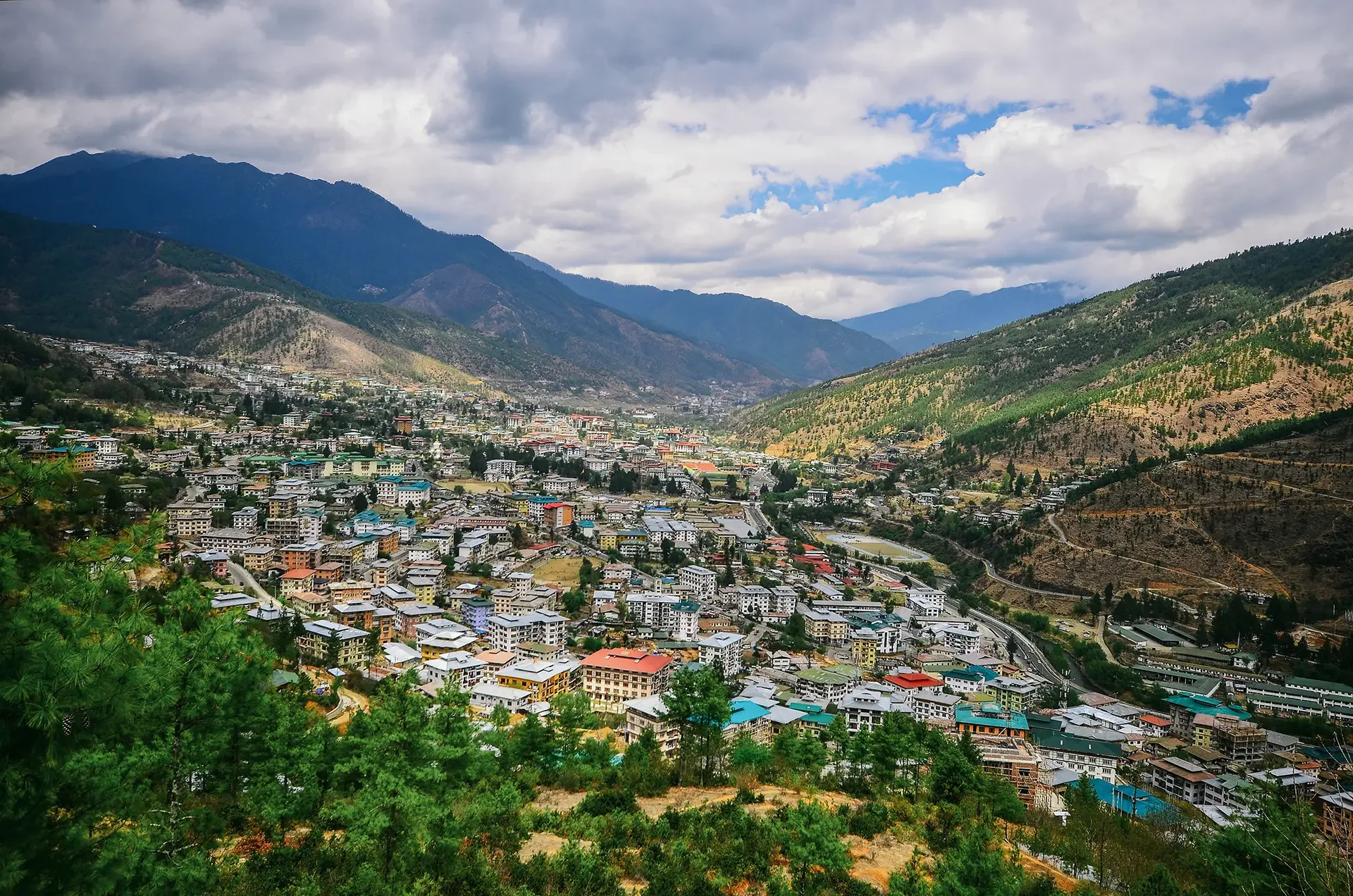 A cityscape in a valley surrounded by green mountains under a partly cloudy sky.