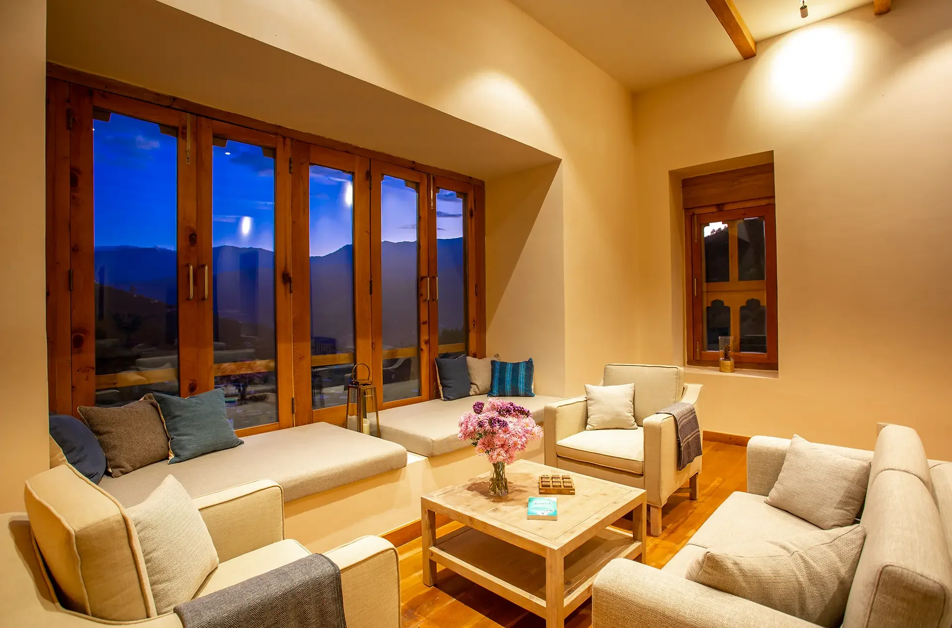 Living room with large wooden framed windows showing a mountain landscape at dusk, beige walls, and cozy seating area with cream-colored sofas and assorted pillows, a wooden coffee table with a flower arrangement, and soft warm lighting.
