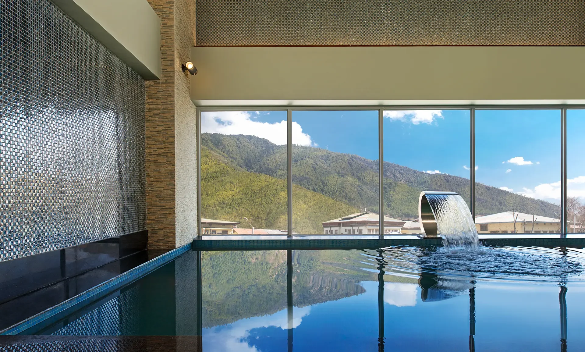 Indoor pool with a large window view of green mountains and blue sky, water feature, textured wall, and modern interior design.