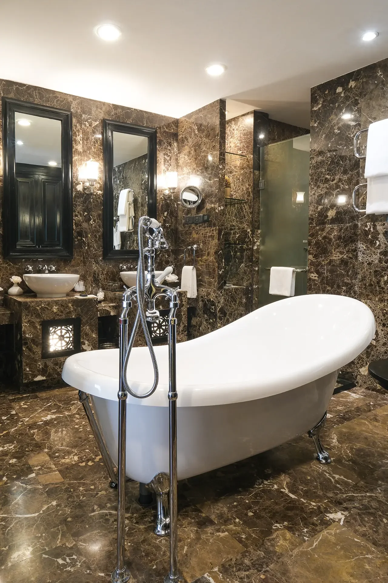 Luxury bathroom with marble walls and floor, freestanding oval bathtub, two sinks with oval mirrors, black framed mirrors, glass shower enclosure, towel rack, and wall-mounted lighting fixtures.