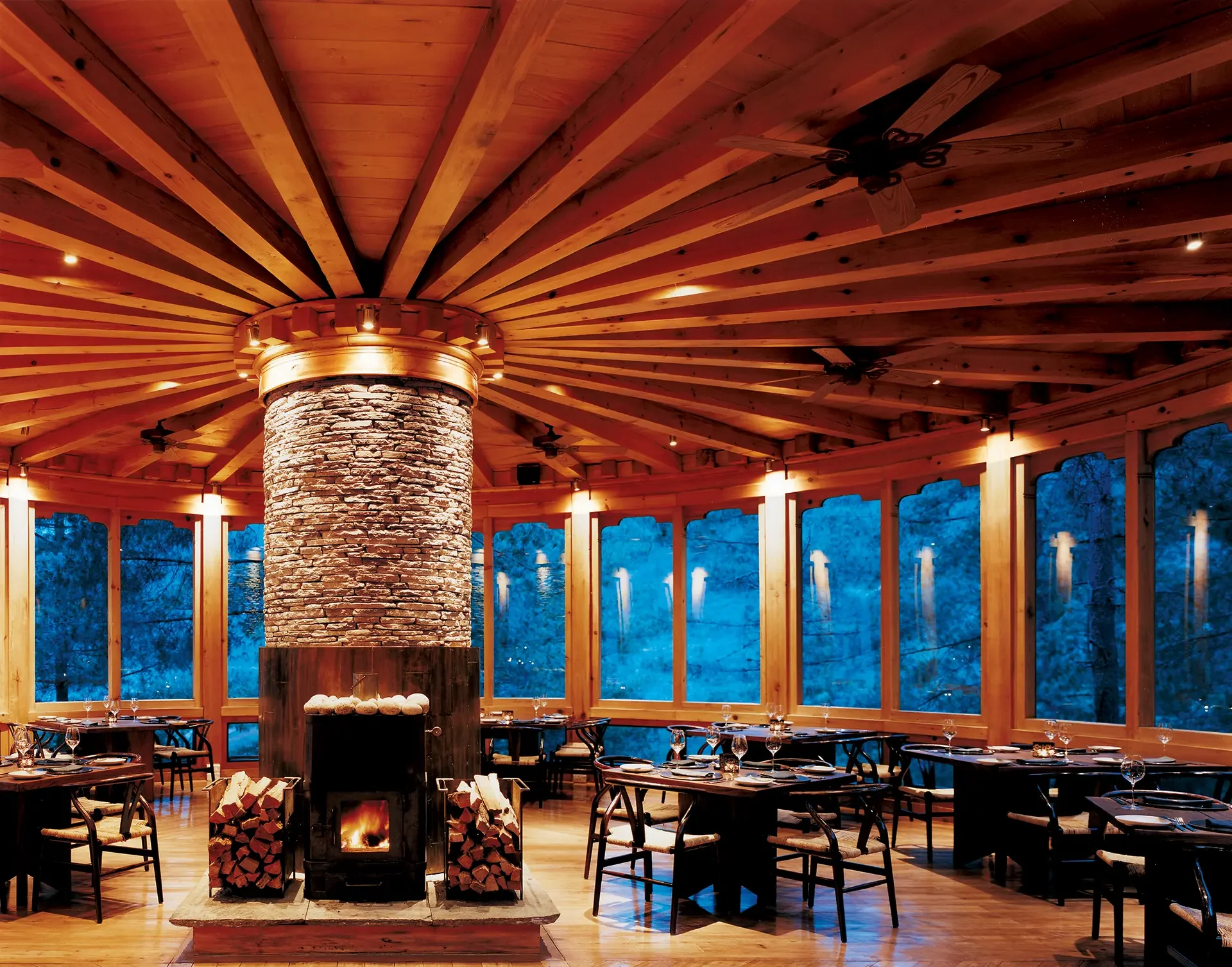Interior of a restaurant featuring a high wooden ceiling with a central stone fireplace, surrounded by tables set with glassware, and large windows showcasing a view of trees at dusk.