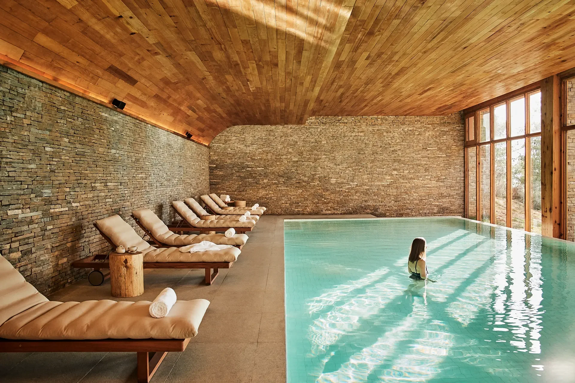 Interior of a spa or pool room with a swimming pool, wooden lounge chairs with rolled towels, large windows, stone and wood walls, and a wooden ceiling.