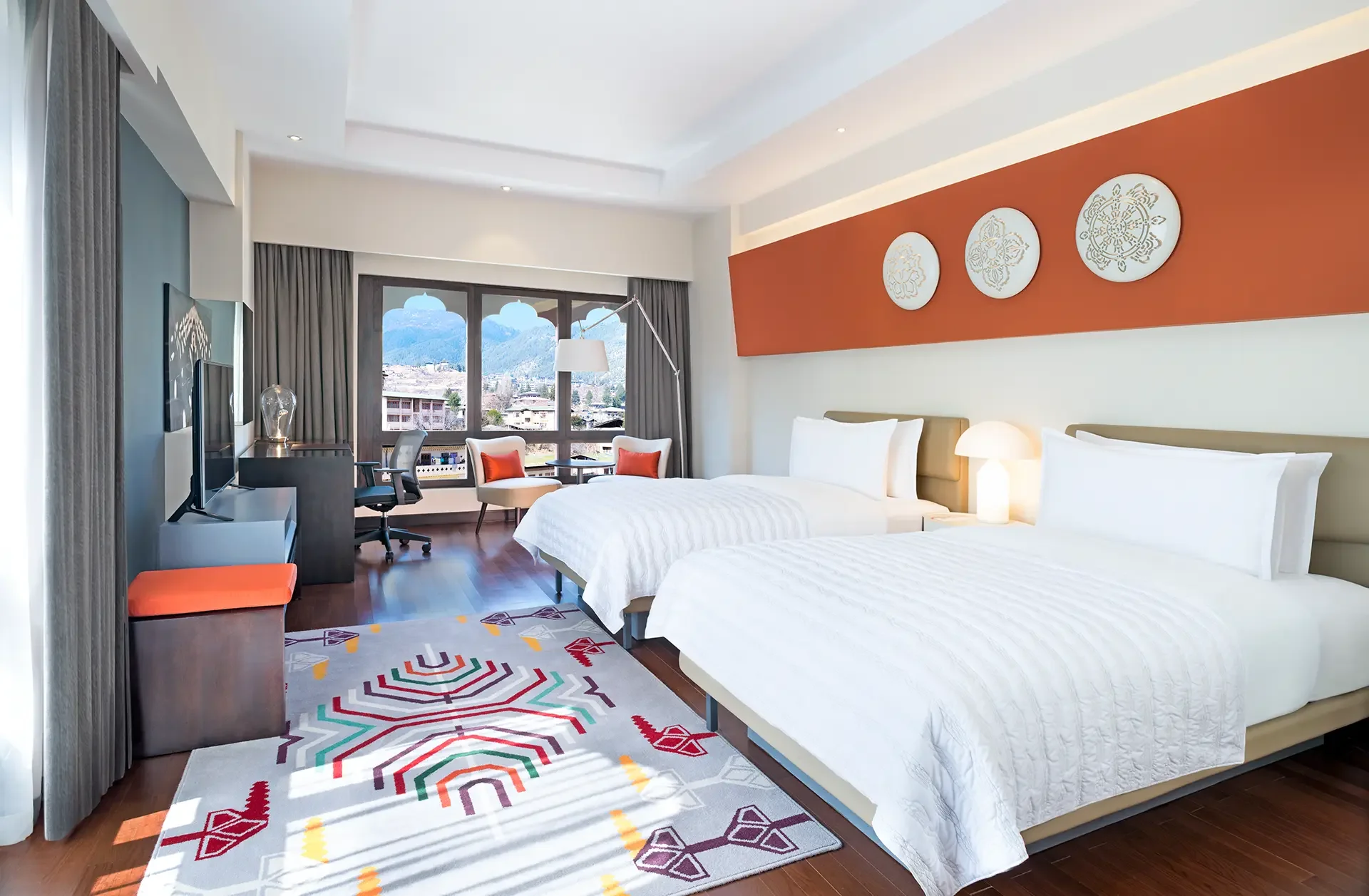 A modern hotel room with two twin beds, white bedding, a bedside lamp, a colorful geometric rug, a window with mountain view, a small sitting area with two chairs and a table, a desk with a chair, and wall art with three circular designs.