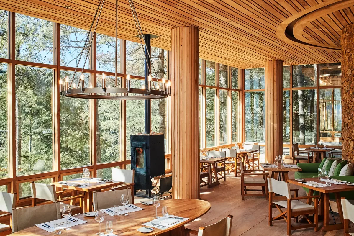 Interior of a cozy restaurant or dining area with large windows, wooden walls, and furniture, including tables set with glasses, plates, and utensils, surrounded by trees visible through the windows.