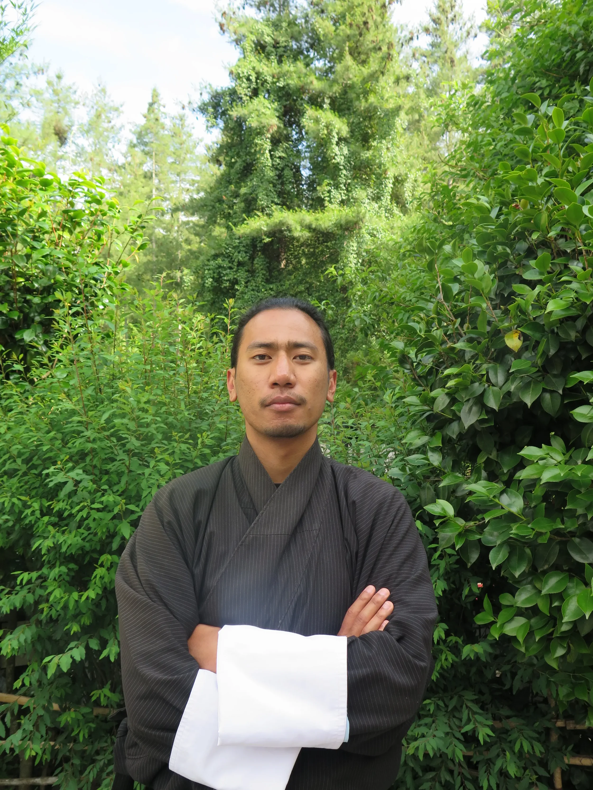 A man wearing traditional Japanese attire with arms crossed standing outdoors among lush green plants and trees.
