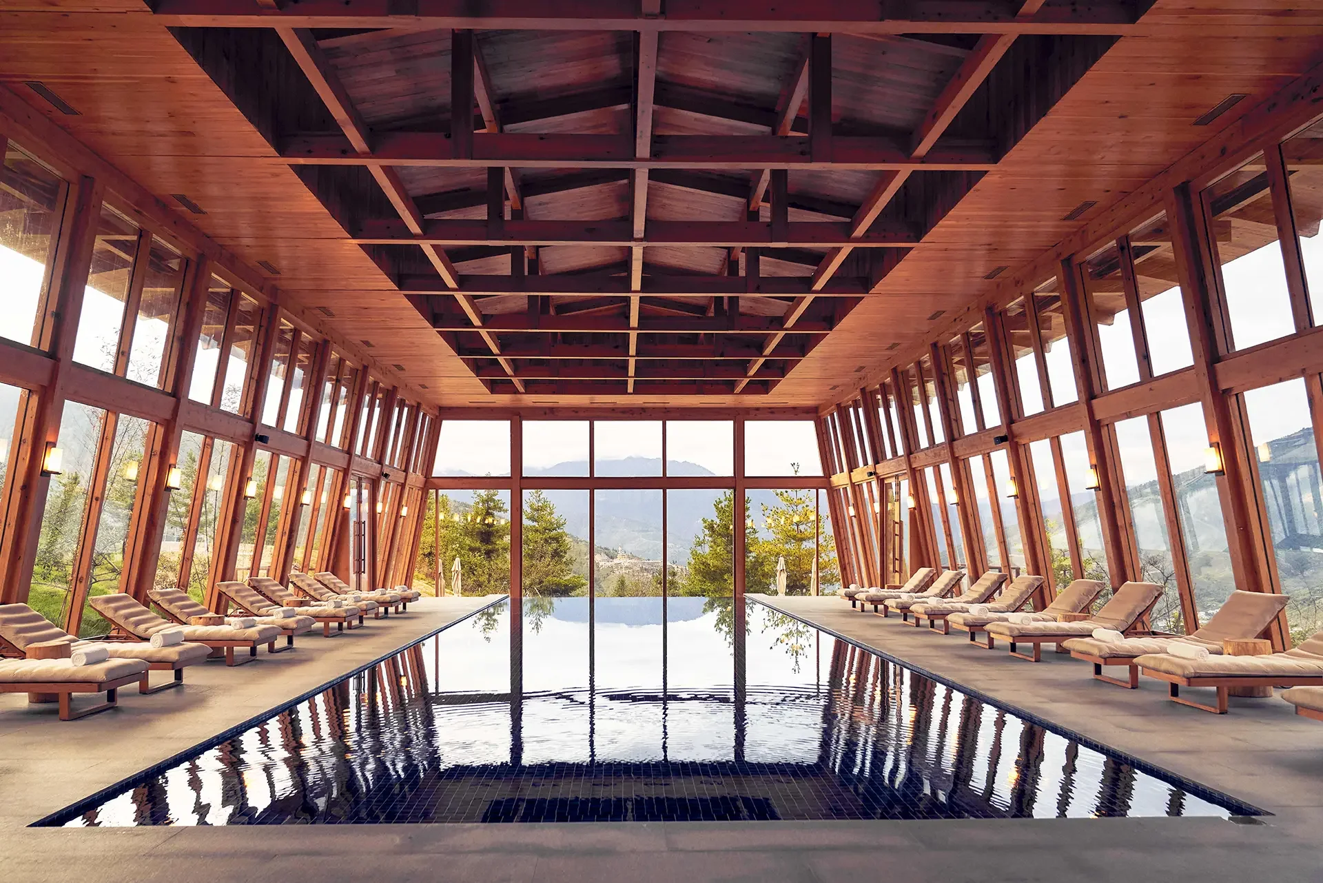 Interior of a wooden spa with large floor-to-ceiling windows and a view of mountains and trees outside. Lounge chairs are arranged along the edge of a rectangular indoor pool with black tiles. The ceiling has exposed wooden beams, adding to the natural, rustic aesthetic.