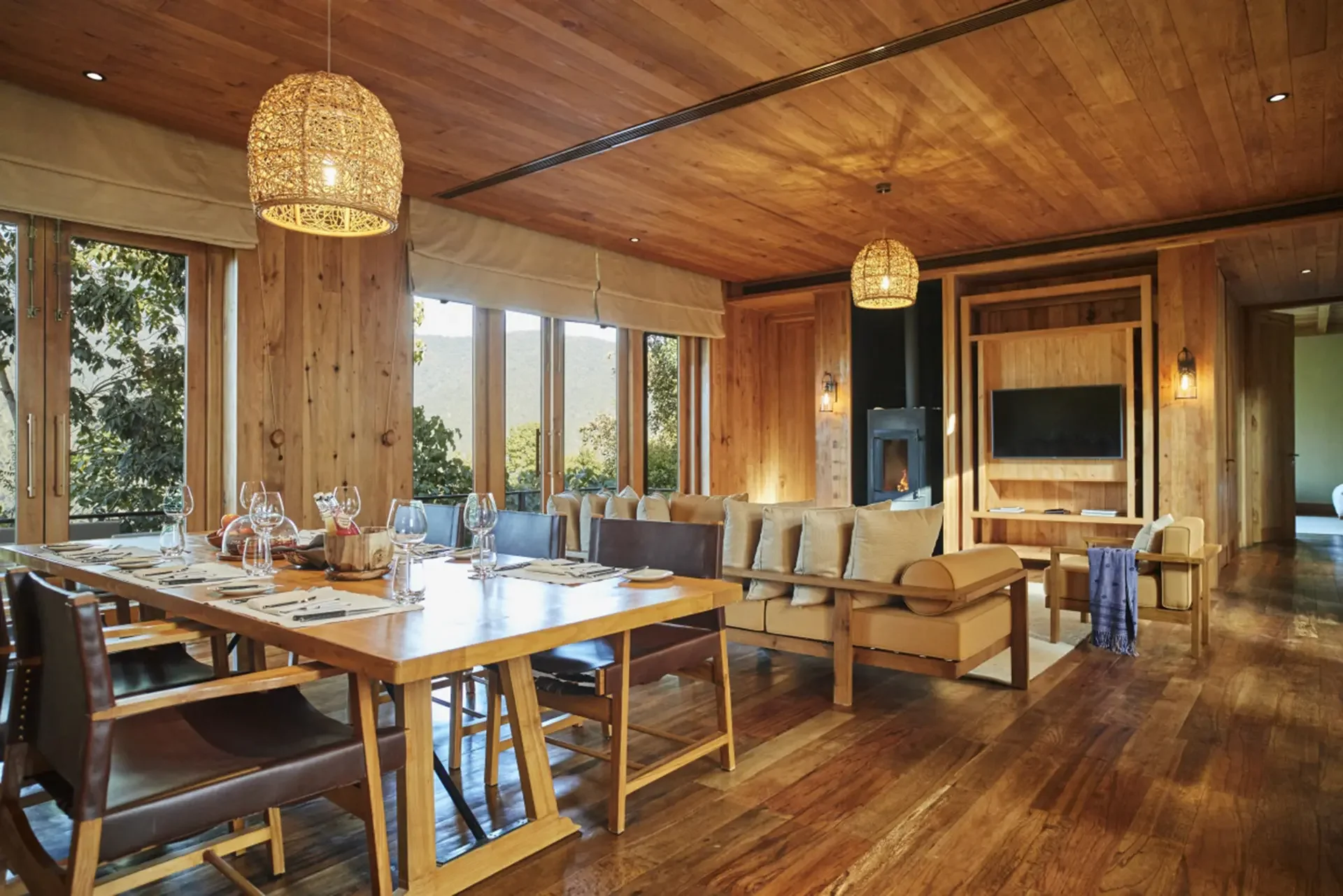 A cozy dining and living room area with wooden paneling, large windows showing trees and mountains outside, pendant lights, a fireplace, a wall-mounted TV, and a long dining table set with glasses and dinnerware.