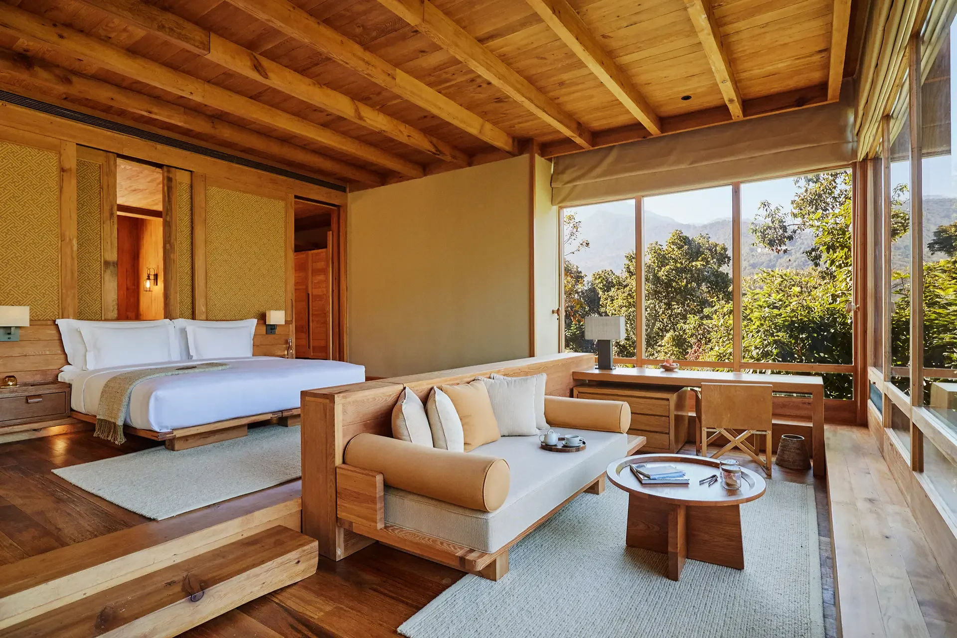 A cozy, modern room with wooden walls, ceiling, and furniture, featuring a large window showing trees and mountains outside, a bed with white linens, a sofa with pillows, and a small coffee table with tea cups.