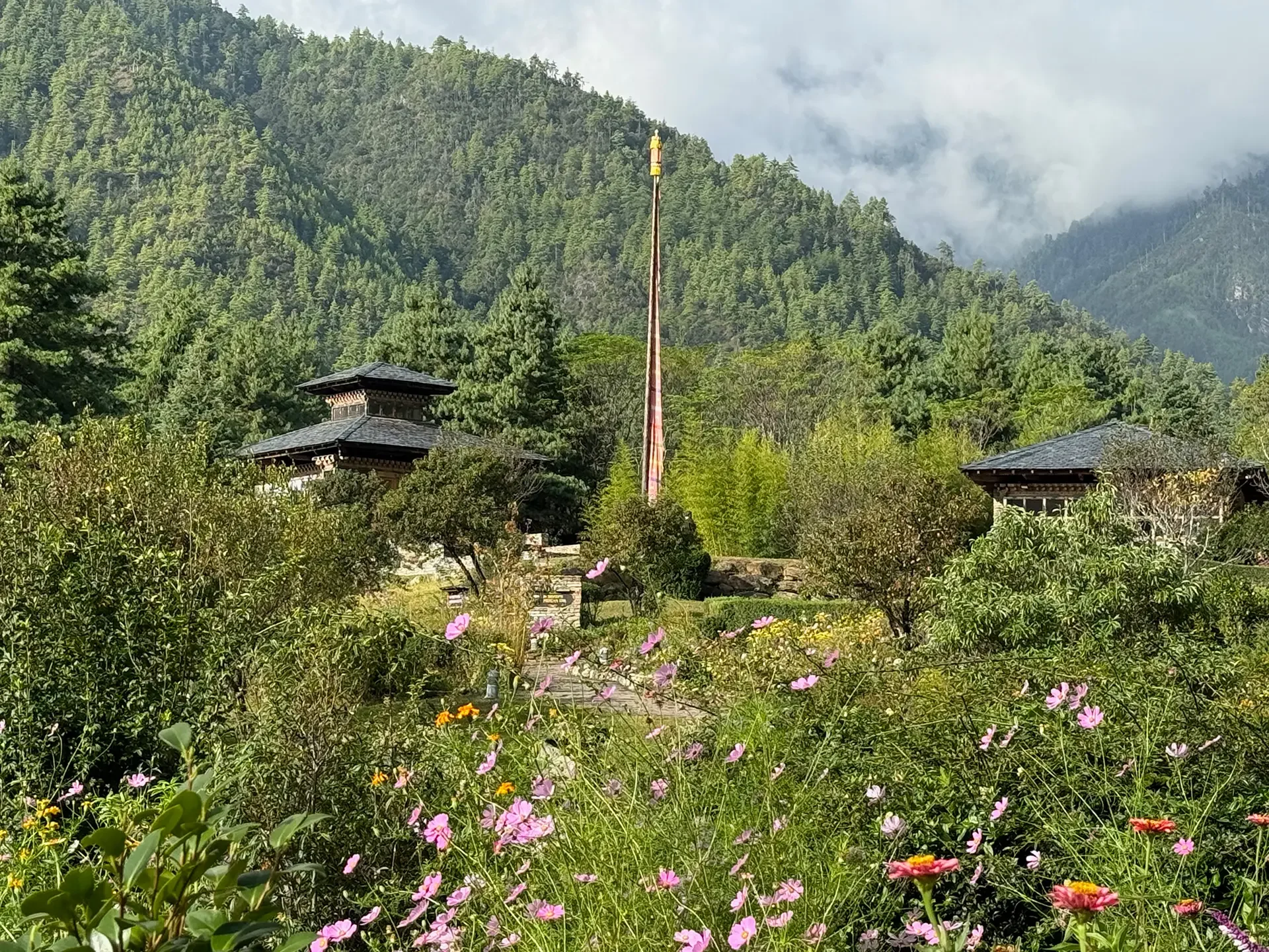 A scenic mountain landscape with dense green pine forests, traditional-style buildings with dark roofs, and a tall flagpole in the foreground. Wildflowers and shrubs fill the lower part of the scene, and misty clouds surround the mountain peaks in the background.