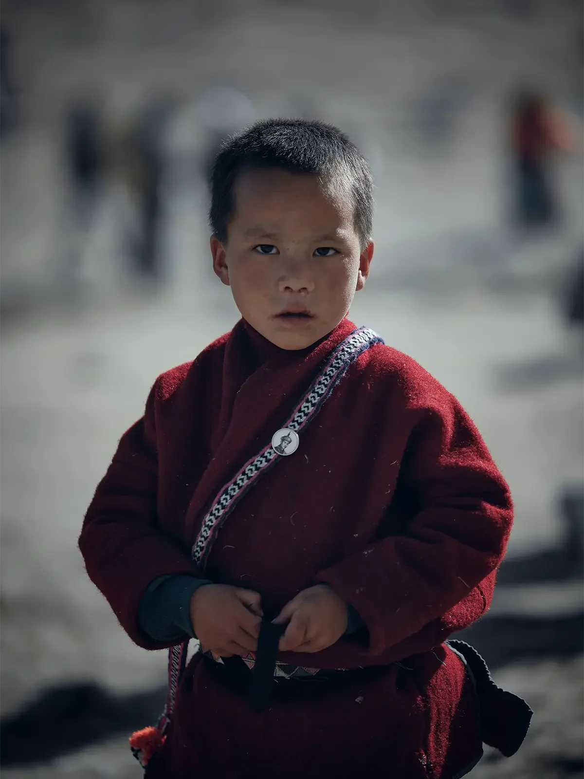 A young boy with short black hair and a serious expression, wearing a red jacket with a patterned strap across his chest, stands outdoors with a blurred background of other people.