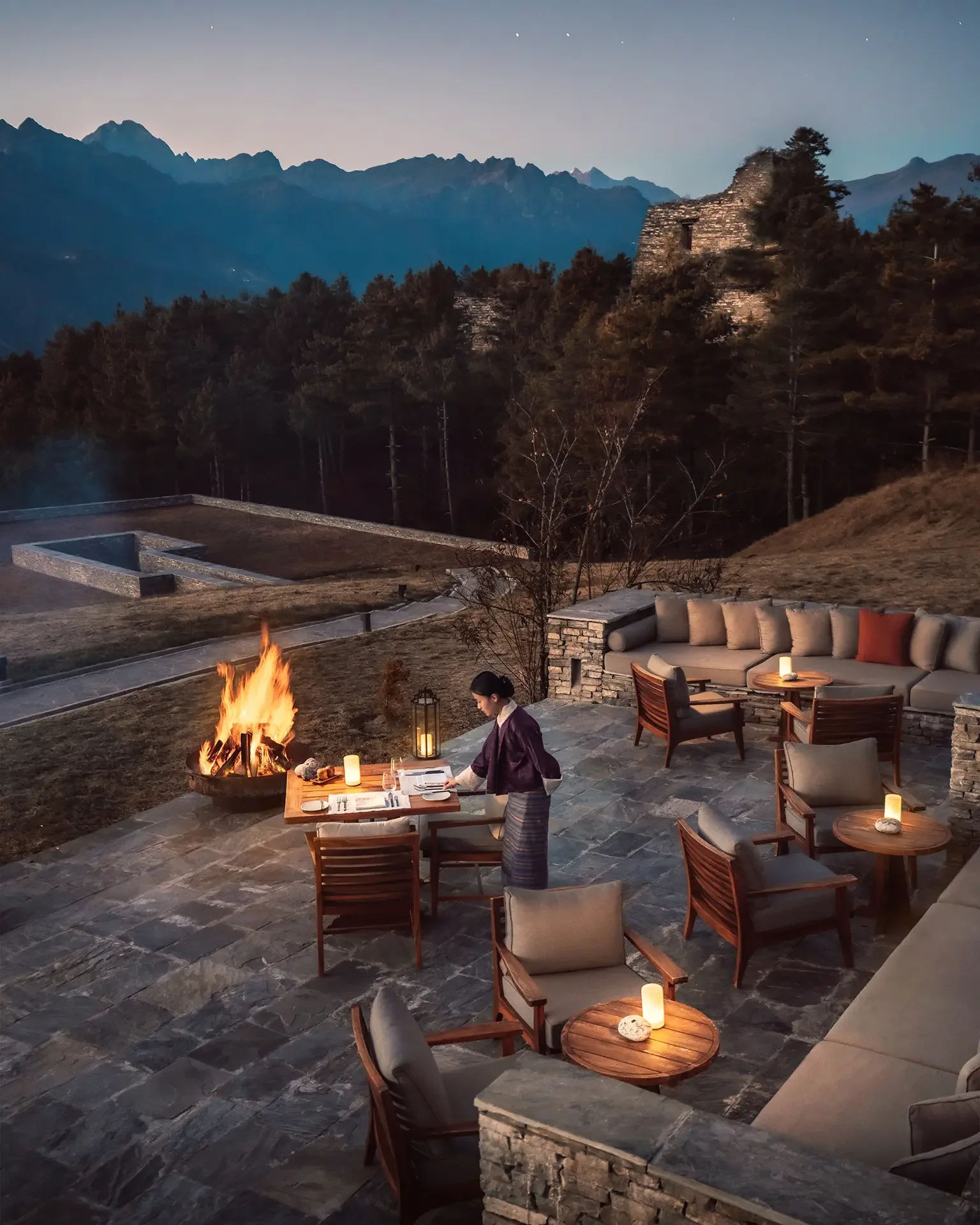 Outdoor lounge area at night with a campfire, dining table, and mountainous background.