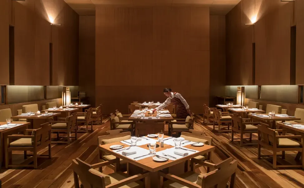 A staff member in a restaurant prepares tables with white placemats, glasses, napkins, and cutlery in an elegant dining area with wooden walls and warm lighting.