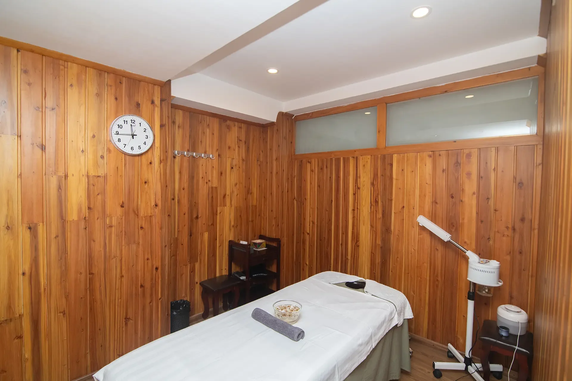 A room with wooden paneled walls and a white ceiling, featuring a wall clock, a corner table, and a massage table with a bowl of supplies, medical equipment, and a lamp.
