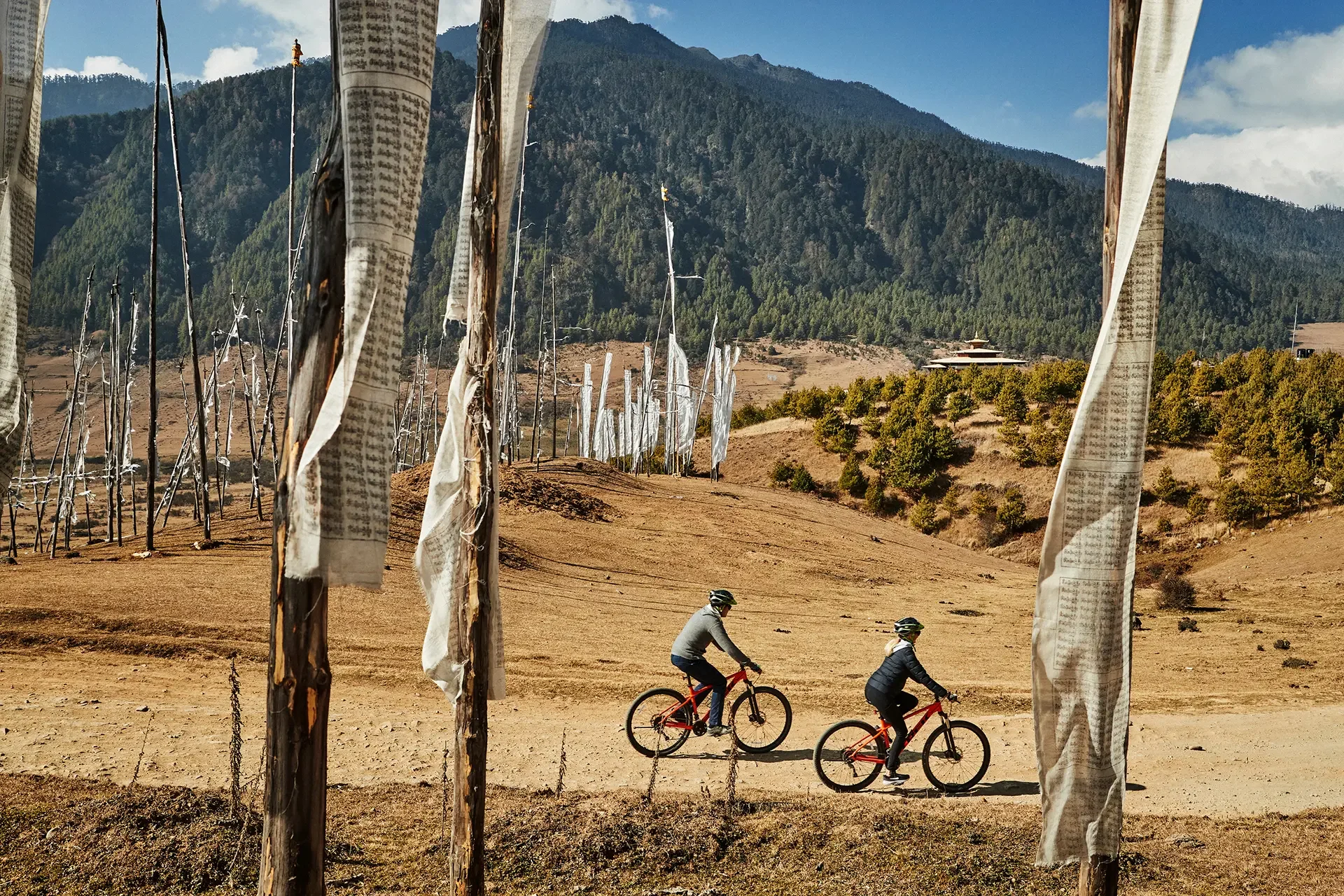 Two children riding bicycles on a dirt path with prayer flags hanging on wooden poles in the foreground. In the background, there are green trees, a hillside, and a mountain under a blue sky with some clouds.