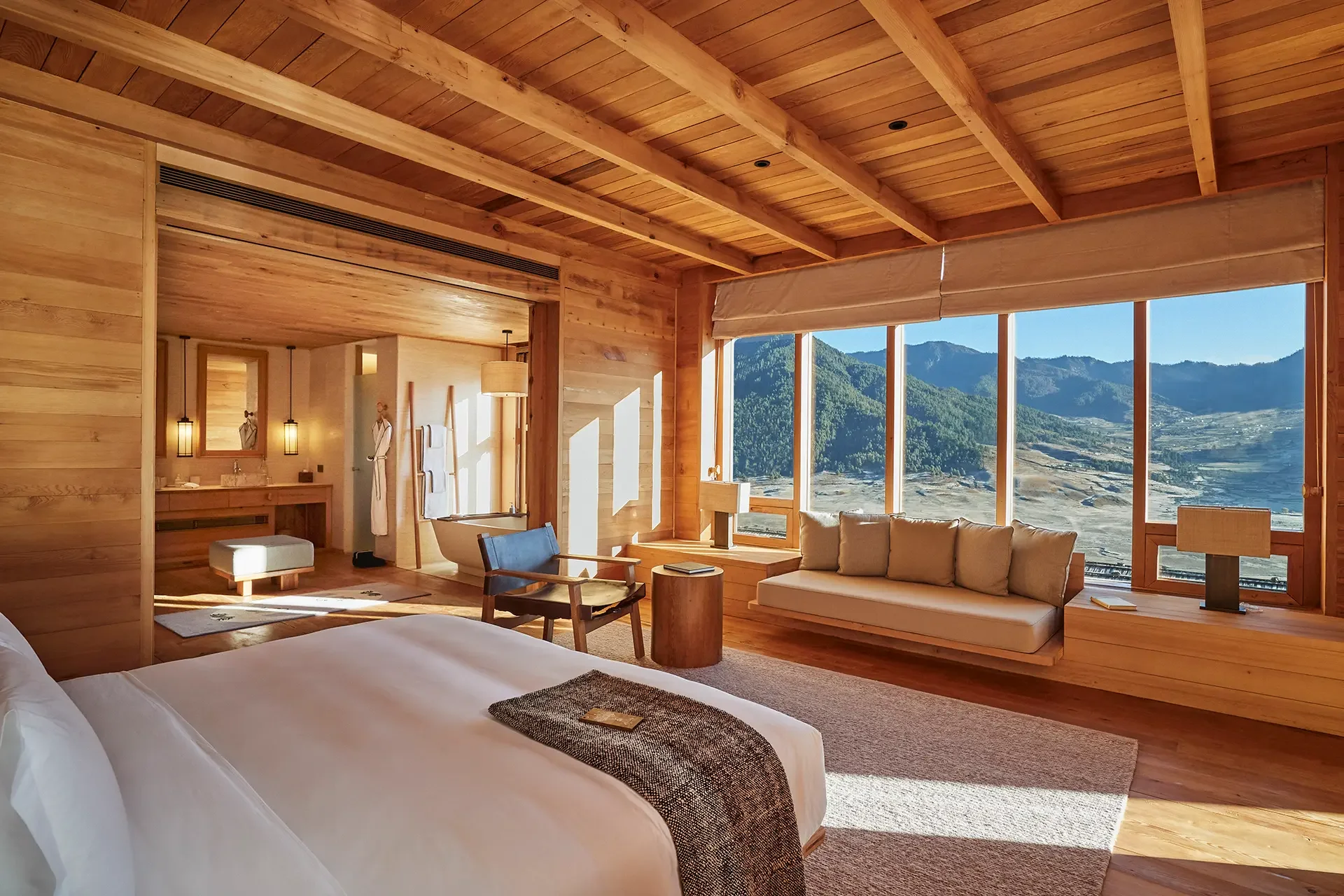 Interior of a wooden lodge room with large windows showcasing mountain views, a bed, sitting area with sofa and chairs, and bathroom area with lights.