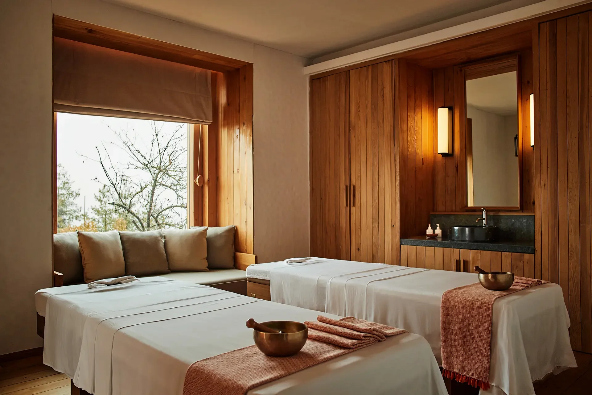 A cozy bedroom with two neatly made beds draped in white linens, topped with folded towels. The room features a large window with a beige shade, wooden wall paneling, and a bathroom area with a sink, mirror, and modern lighting. A brass bowl and a small tray with bottles are on the beds.