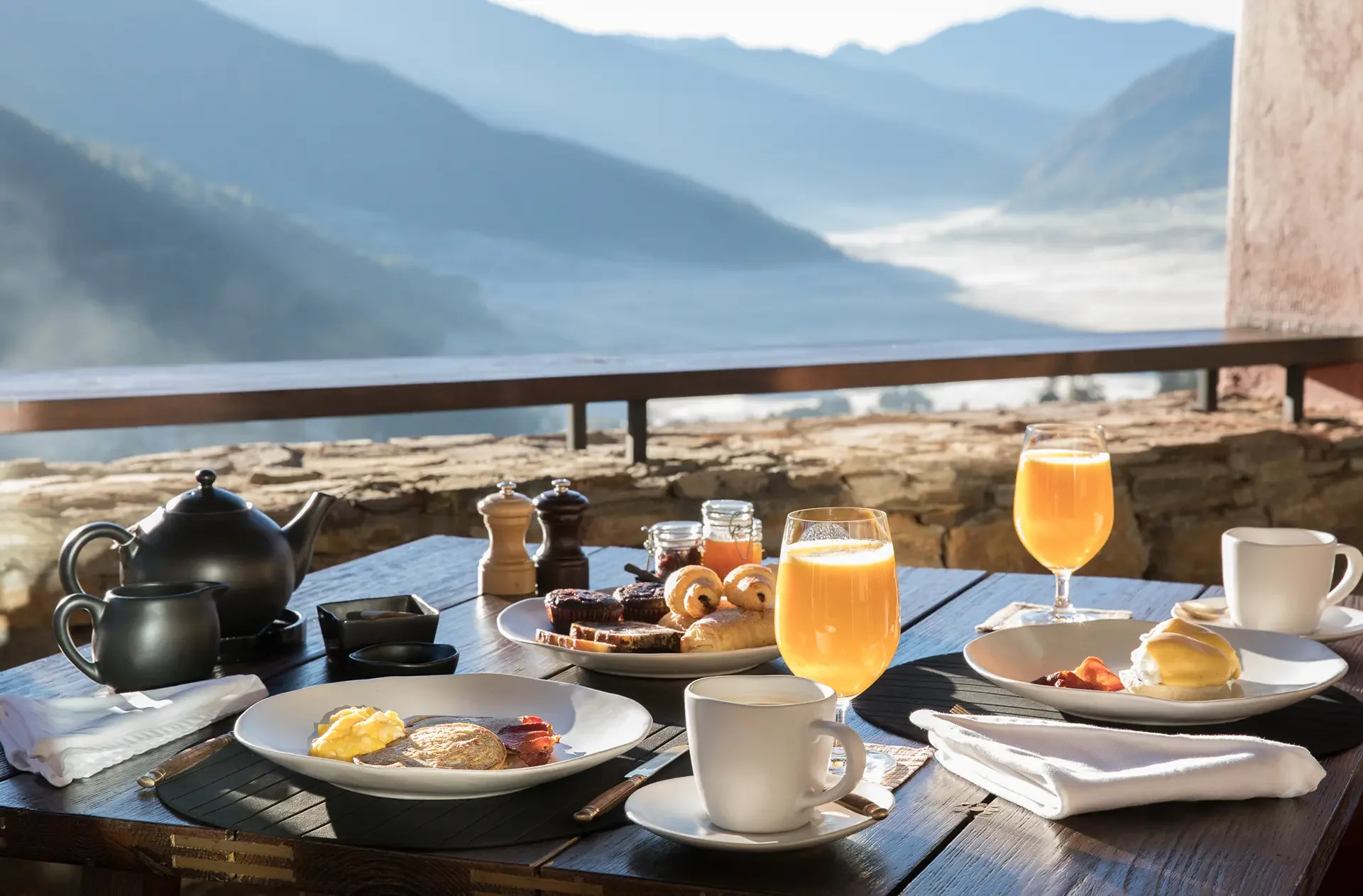 Breakfast spread on a wooden table with coffee, orange juice, muffins, pastries, and egg dishes, overlooking mountainous scenery through a large window.