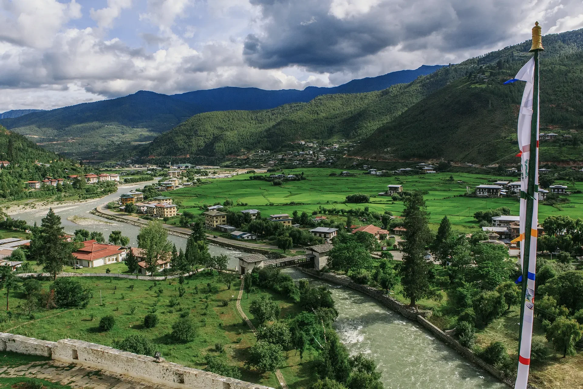 Scenic view of a river flowing through a lush green valley with houses, surrounded by mountains under a cloudy sky, with a flagpole on the right side.