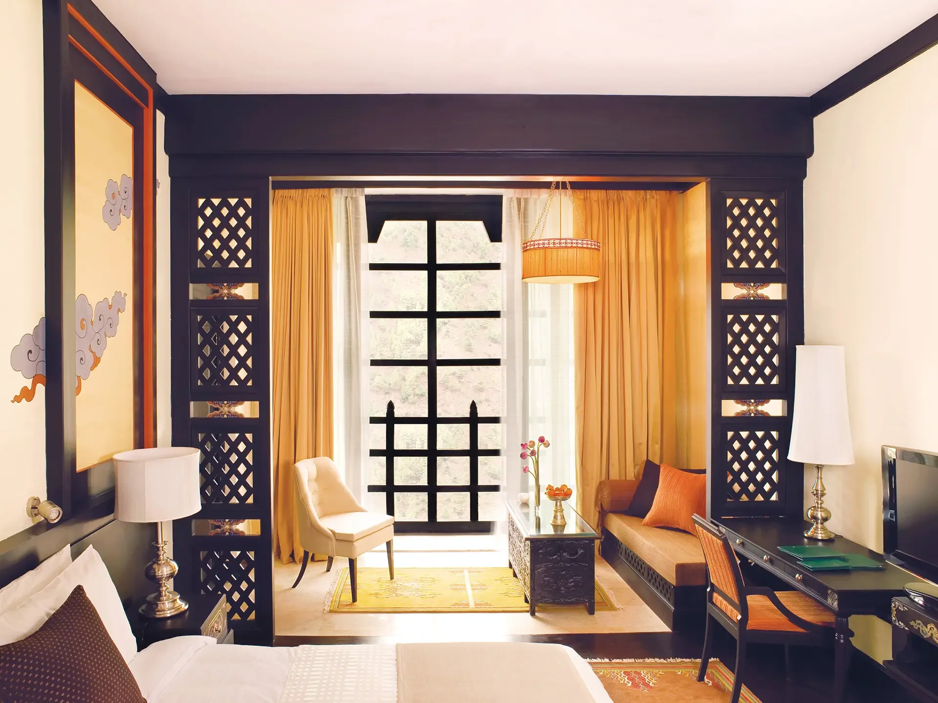 A hotel room with a bed, side table, and lamps on each side. A sitting area with a yellow chair and a brown sofa with orange cushions, a glass-top coffee table, and a large window with yellow curtains. The window has black grid panes and shows a view of greenery outside.