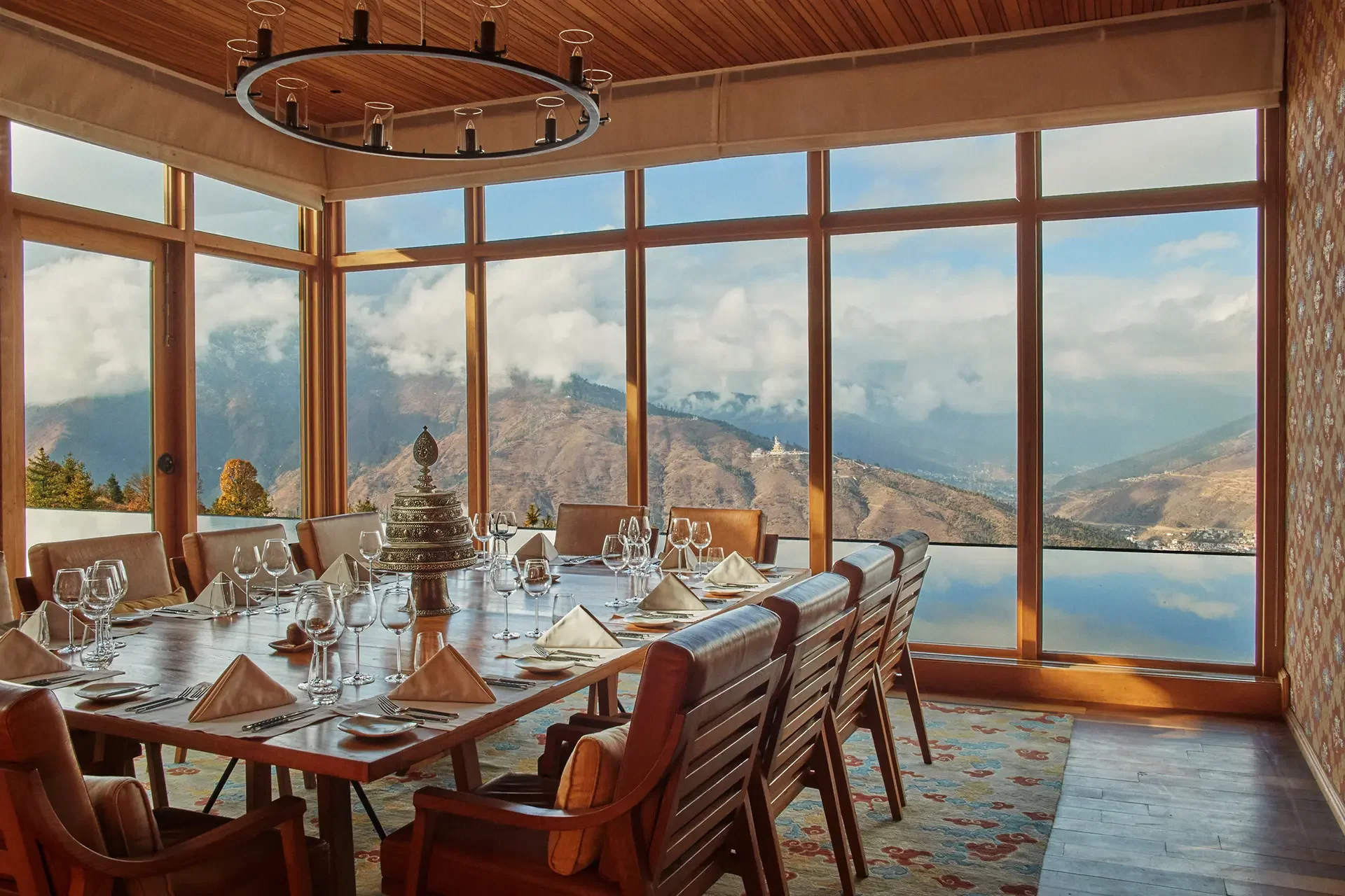 A dining room with a large wooden table set with wine glasses, folded napkins, and cutlery, surrounded by brown leather chairs, and large windows offering a mountain and cloudy sky view.