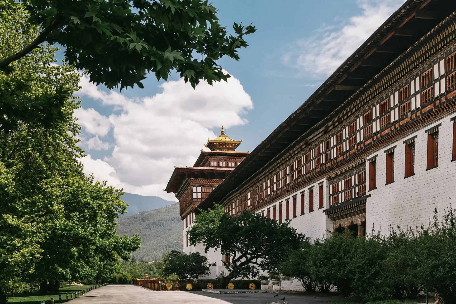 A traditional multi-story building with ornate wooden windows and a golden rooftop, surrounded by green trees and set against a backdrop of mountains and a partly cloudy sky.