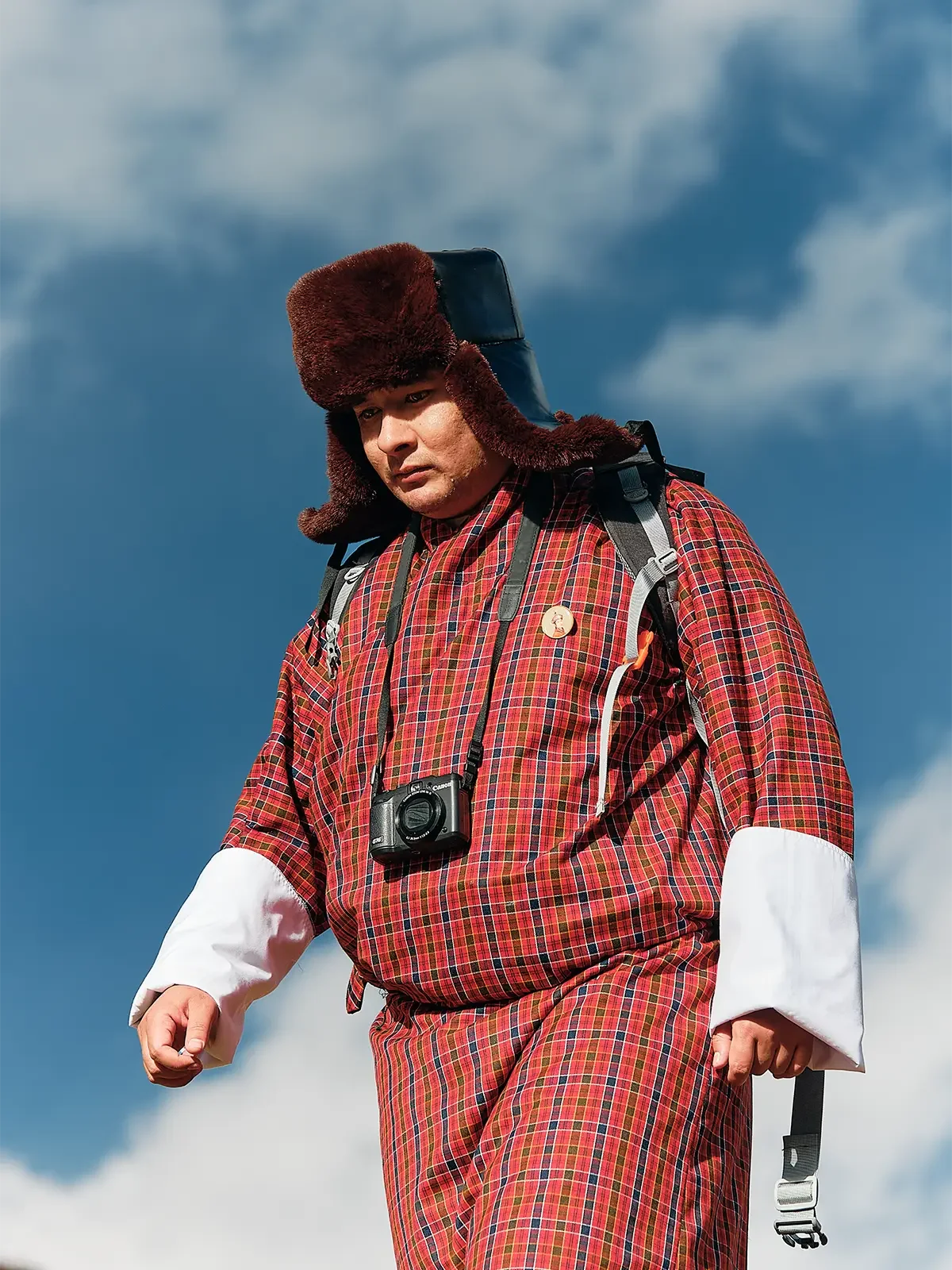 A man wearing a red plaid jacket and pants, a thick fur hat, and a camera around his neck, stands outdoors against a cloudy sky.