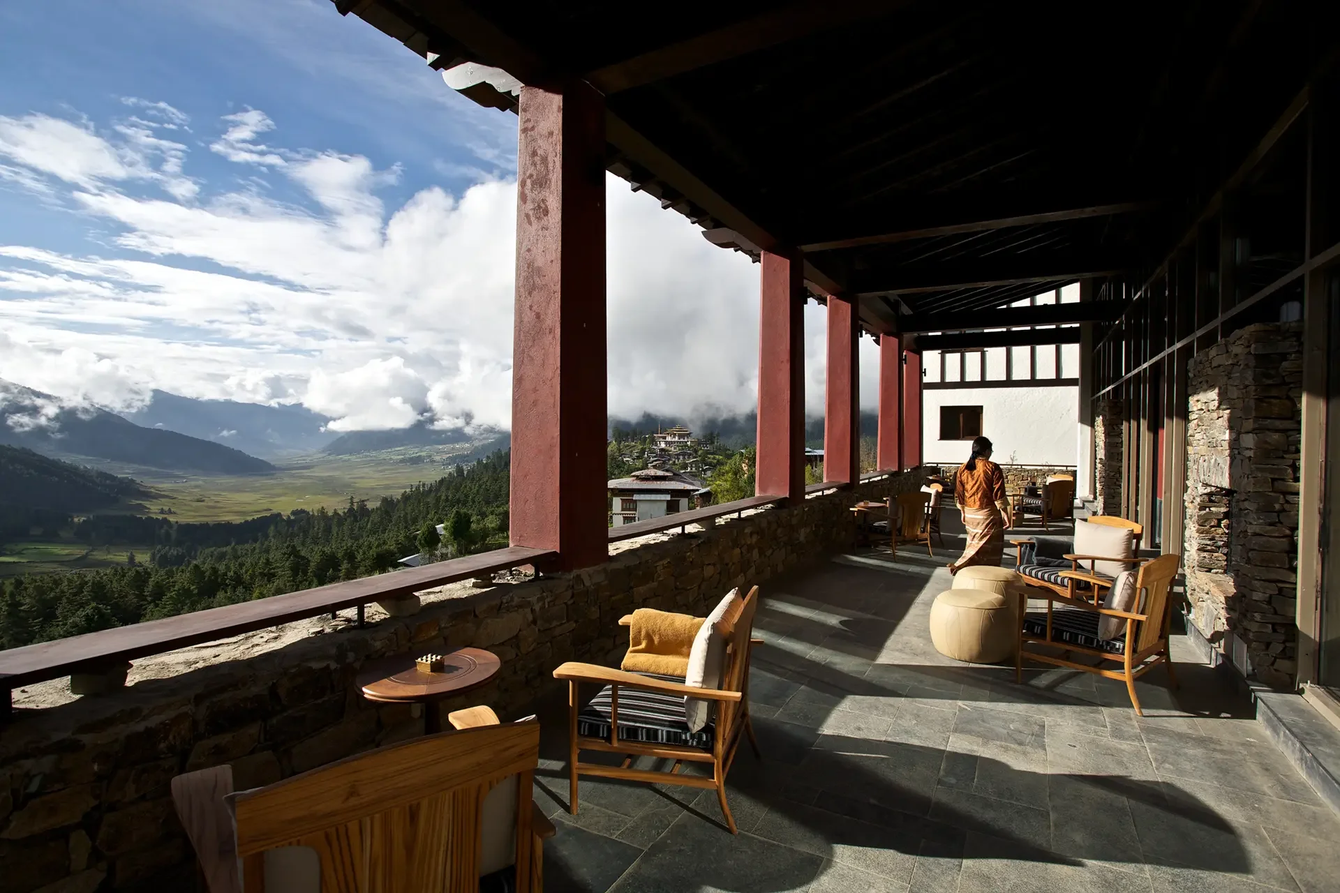 A spacious balcony with outdoor seating overlooking a scenic mountainous landscape, with a person wearing traditional clothing walking through the area.