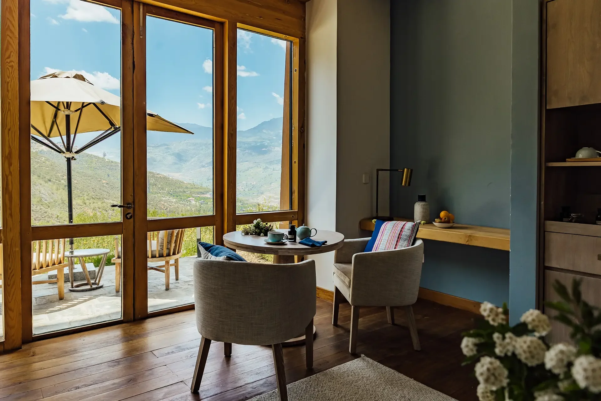Interior view of a cozy dining area with a round table set for tea or coffee, surrounded by chairs, next to large glass doors open to a patio with outdoor chairs and an umbrella, overlooking a mountainous landscape under a blue sky with scattered clouds.