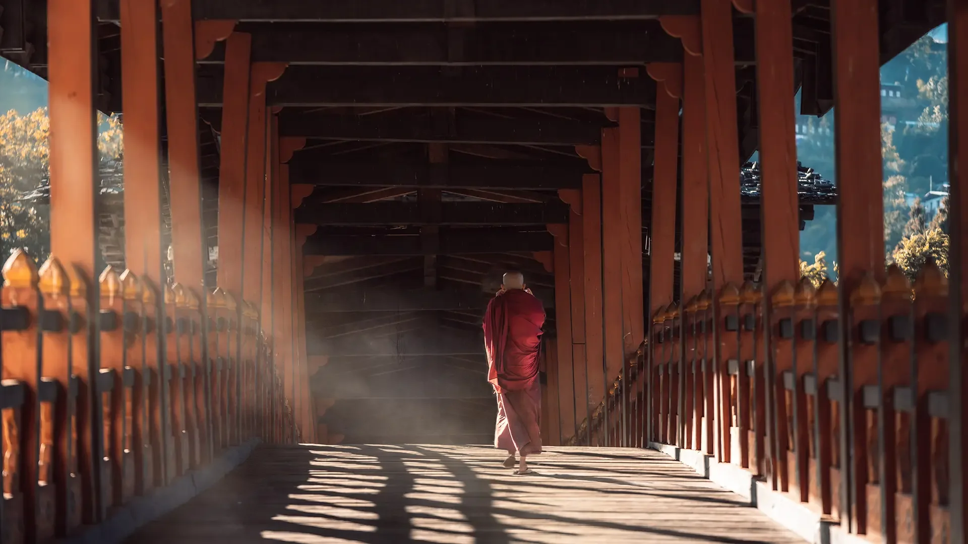 A person dressed in a red robe walking under a wooden bridge with intricate railings, with sunlight casting shadows on the bridge deck and mountains in the background.