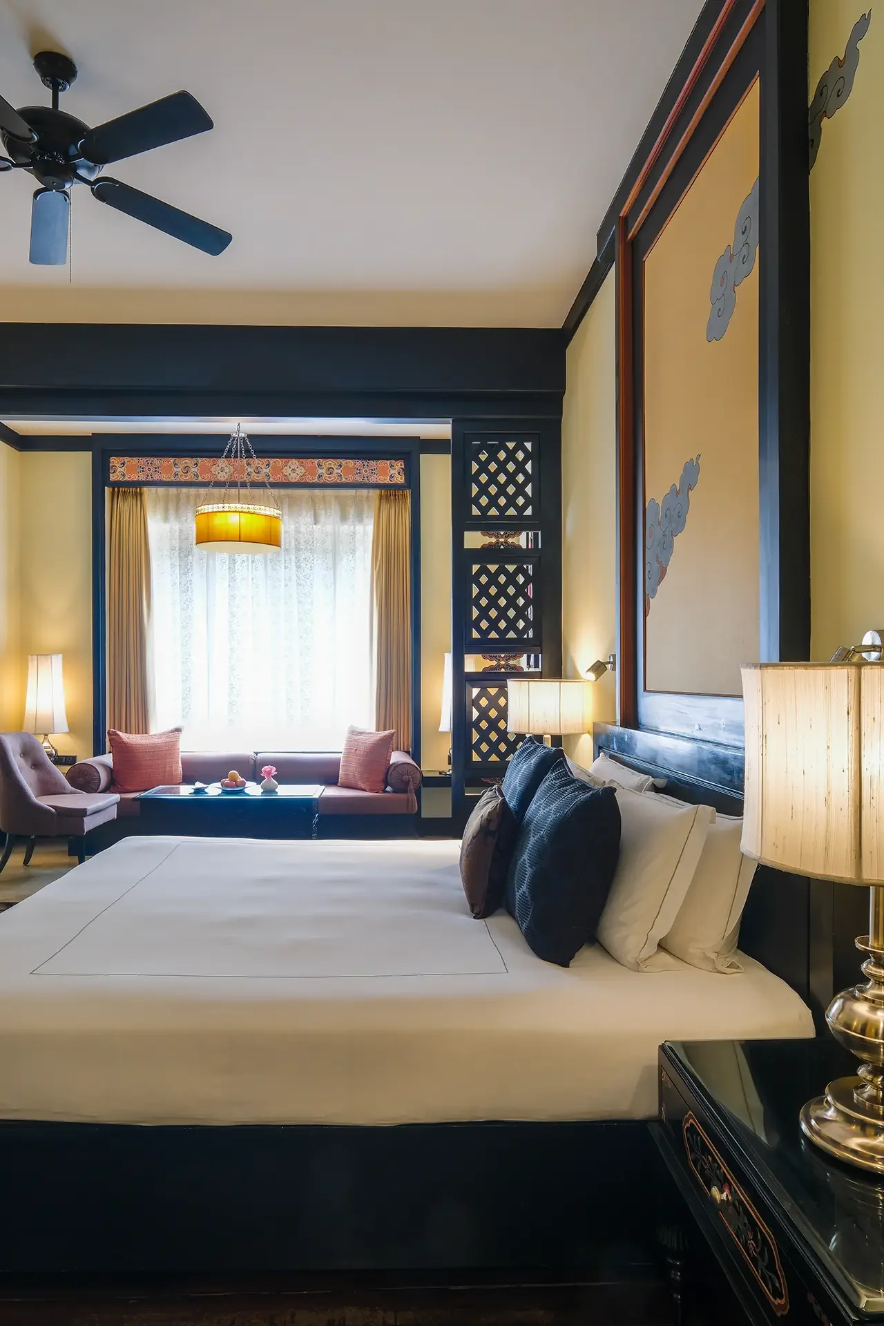 A luxurious hotel room with a large bed, black and white pillows, a bedside lamp, and a black side table. In the background, a sitting area with a pink sofa, armchair, coffee table, and decorative pillows, with a window and curtains, a hanging ceiling light, and a wall with traditional Asian artwork.