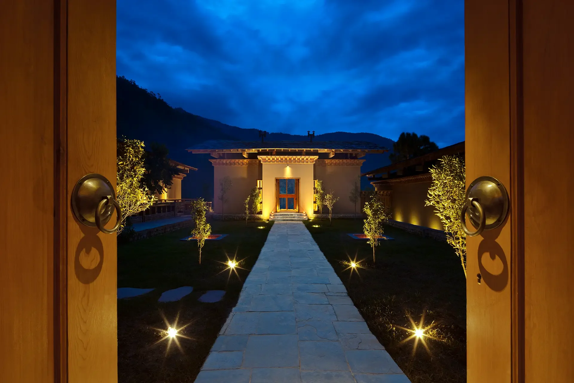 View from open wooden door looking into a well-lit yard with a paved pathway leading to a house, surrounded by neatly trimmed trees and landscaped garden at night, with mountains and a cloudy sky in the background.