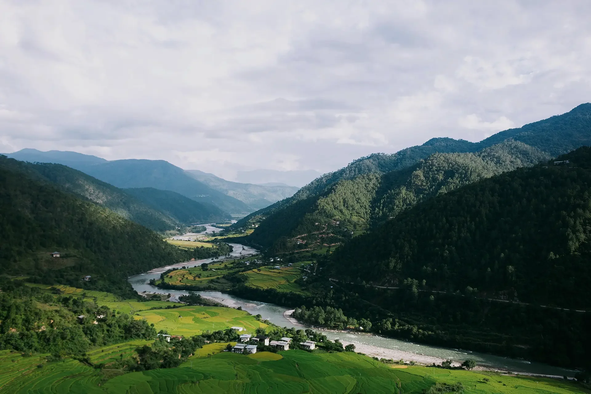 Scenic view of lush green mountains with a river flowing through a valley, patchwork of farmland, and a cloudy sky overhead.