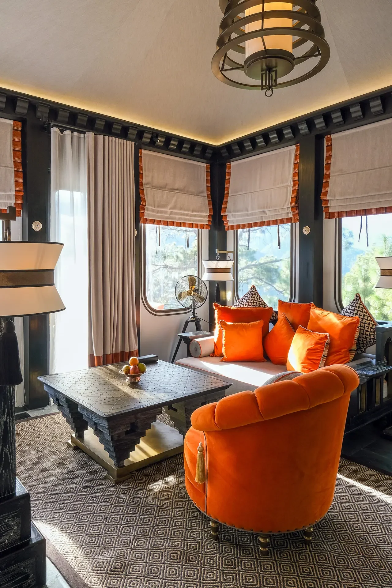 Living room with black frame windows, beige Roman shades, beige curtains, orange and patterned cushions, orange plush armchair, black-trimmed table with fruit bowl, patterned carpet, black pillar, ceiling light fixture.