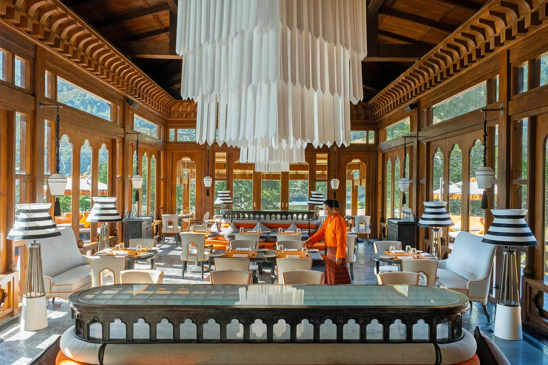 A bright, airy restaurant with wooden walls and ceiling, large windows, and modern decor. A white, tiered chandelier hangs from the center. A server in orange is preparing a table.
