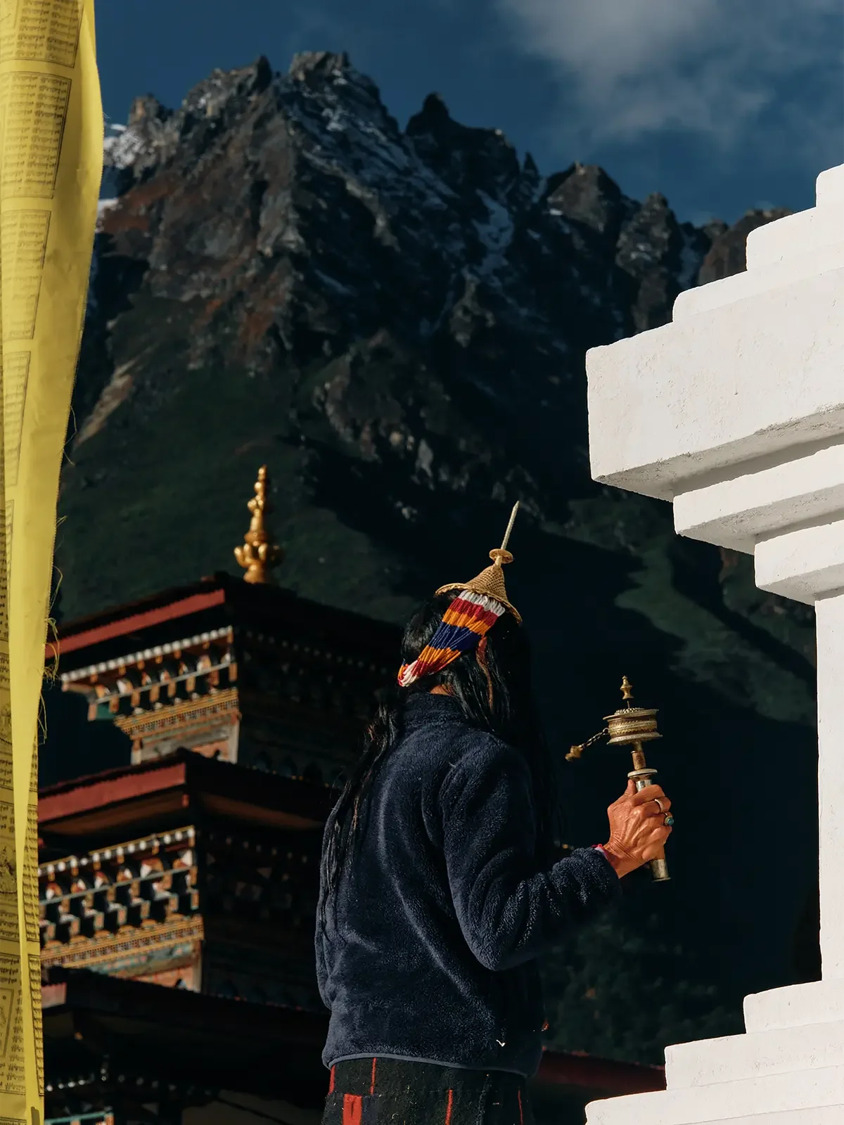 A woman wearing a colorful headscarf and black jacket, holding a prayer wheel, standing in front of a temple with intricate architecture, with a mountain range in the background.