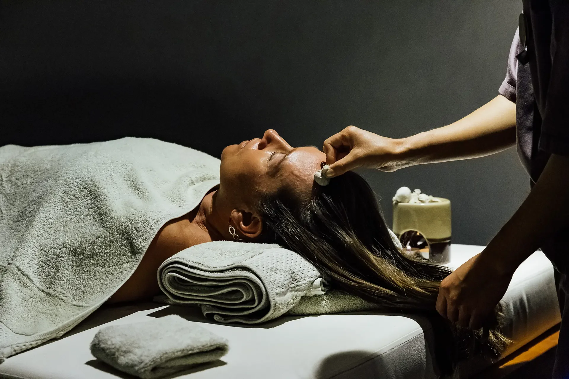 A woman receiving a facial treatment while lying on a spa bed with a towel and a rolled blanket, as a skincare professional applies treatment to her face in a spa setting.