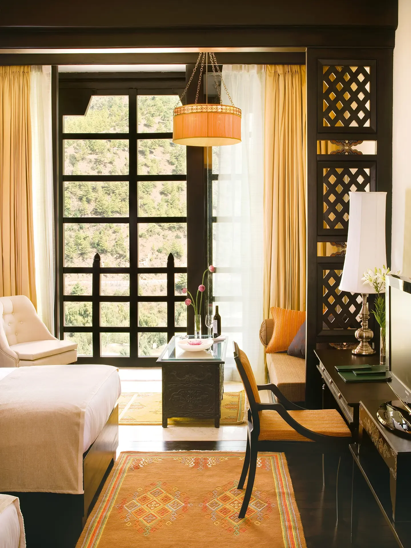 Living room with a large window showing trees outside, beige and yellow curtains, a beige sofa, a black side table with a pink flower arrangement, a black desk with a lamp and decorative items, and a patterned orange rug.