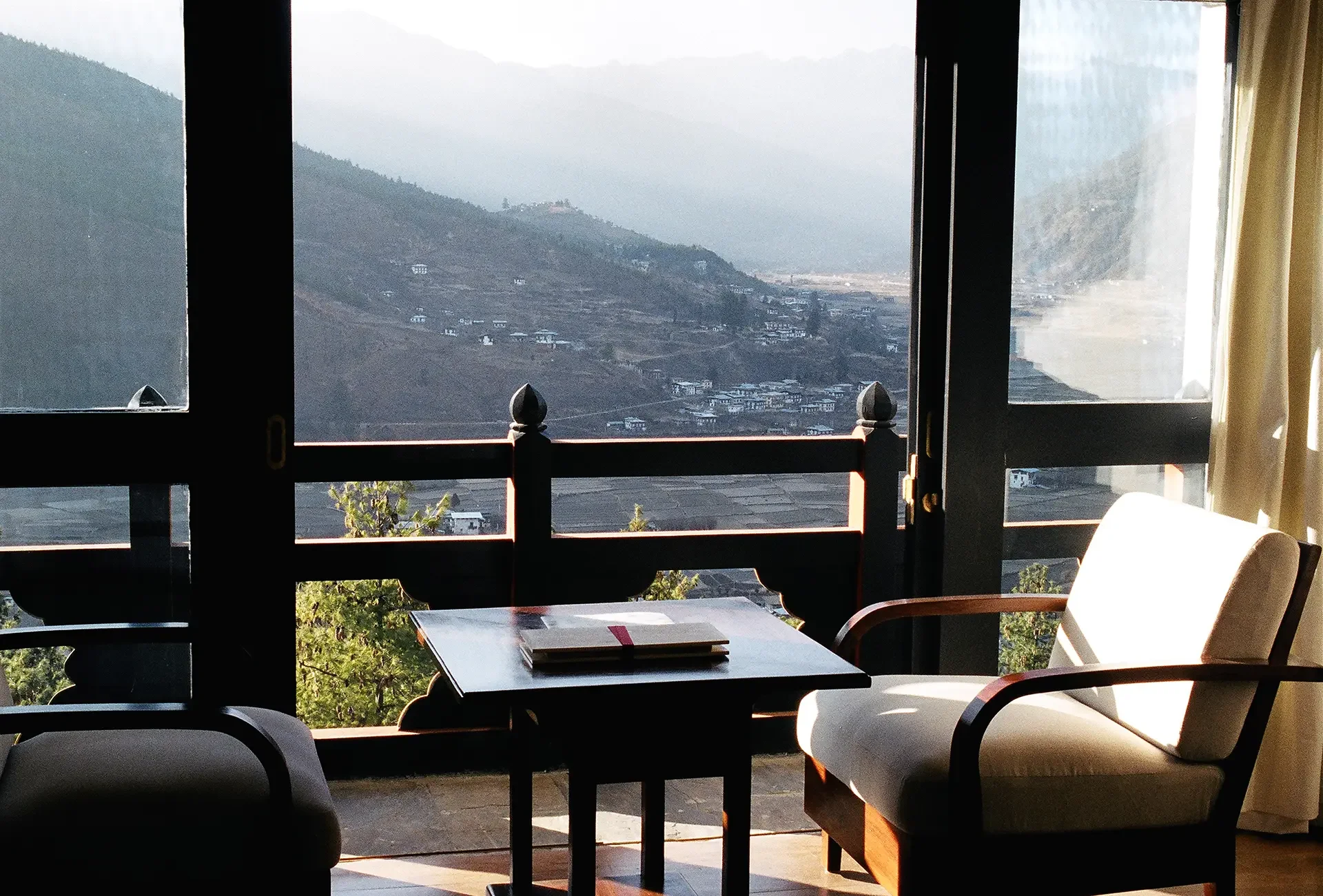 Interior of a room with large windows overlooking a mountainous landscape with small buildings, a table with a menu or brochure, and chairs.
