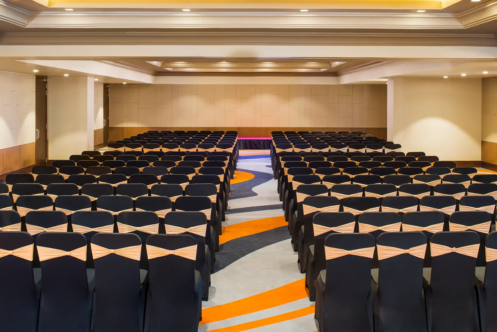 Empty conference room with rows of black chairs decorated with peach-colored bows, arranged in front of a stage with a red and purple curtain, with beige walls and a ceiling with recessed lighting.