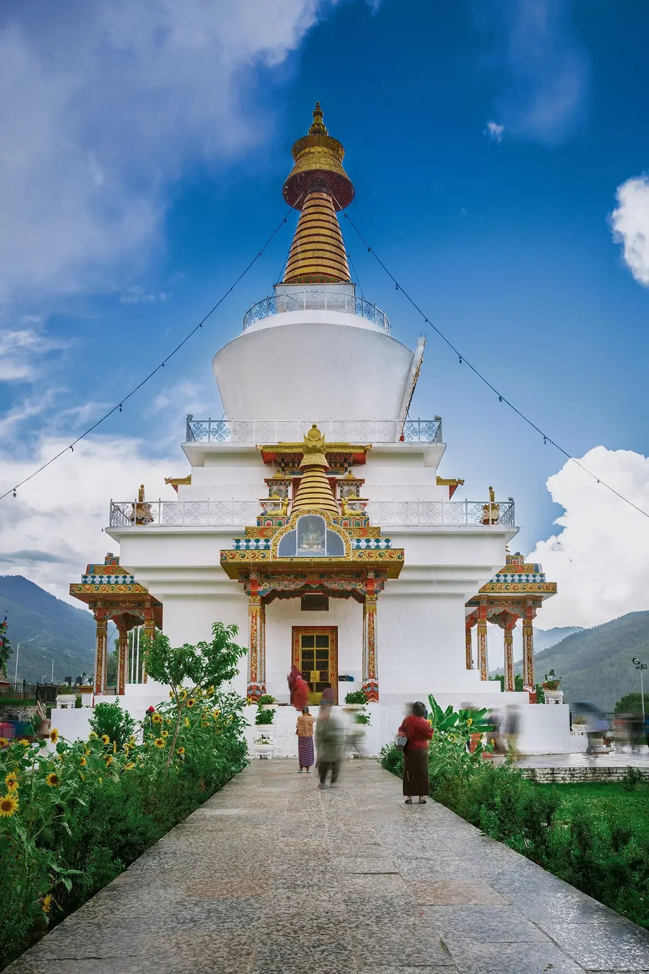 A white Buddhist stupa with colorful decorative details and a golden spire, set against a partly cloudy sky with mountains in the background. There are people walking towards the entrance, with a garden of sunflowers and green plants in the foreground.