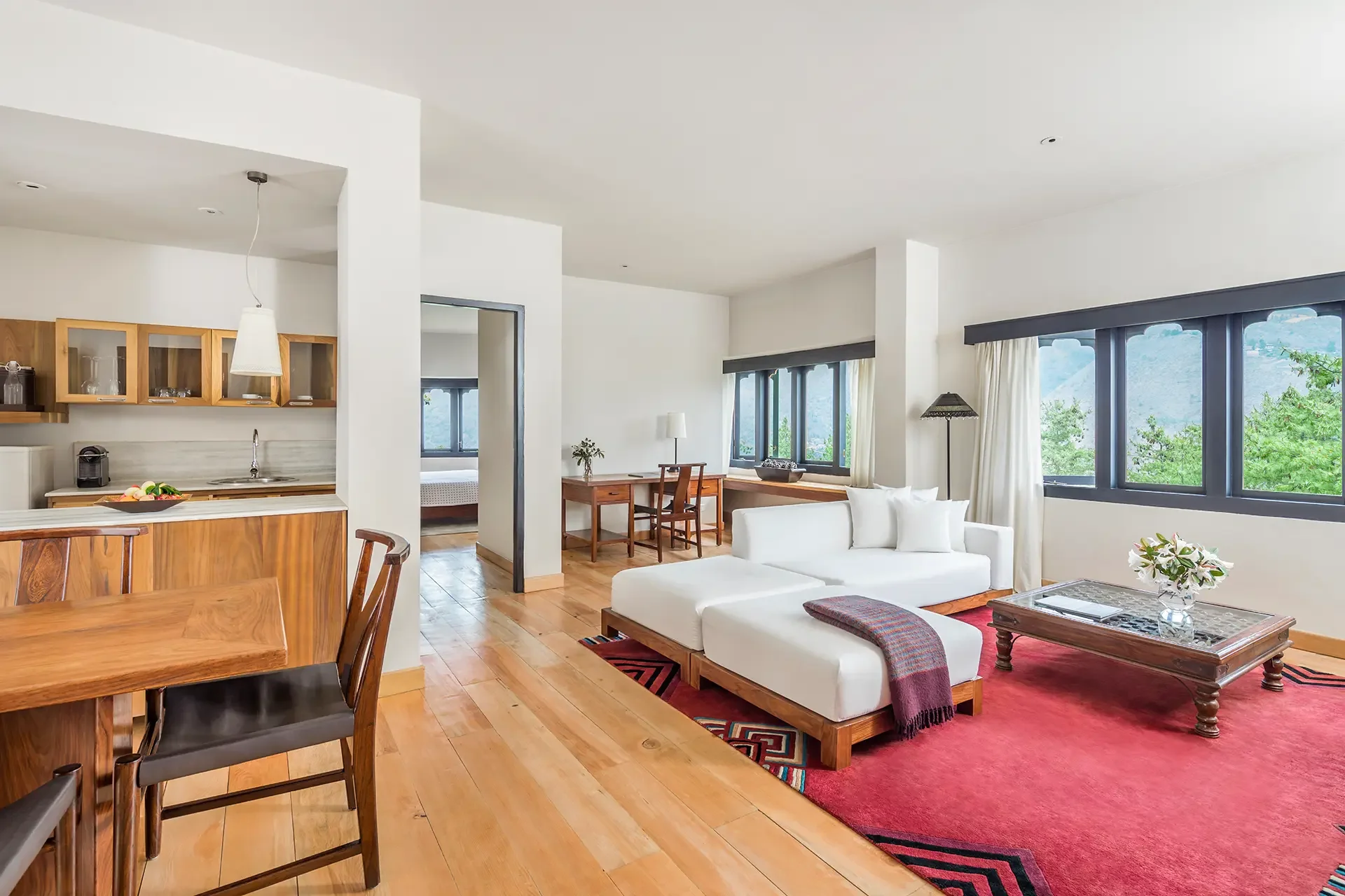 Living room with white sofas, wooden coffee table with flowers, large windows with scenic mountain view, hardwood floors, and a kitchen area in the background.