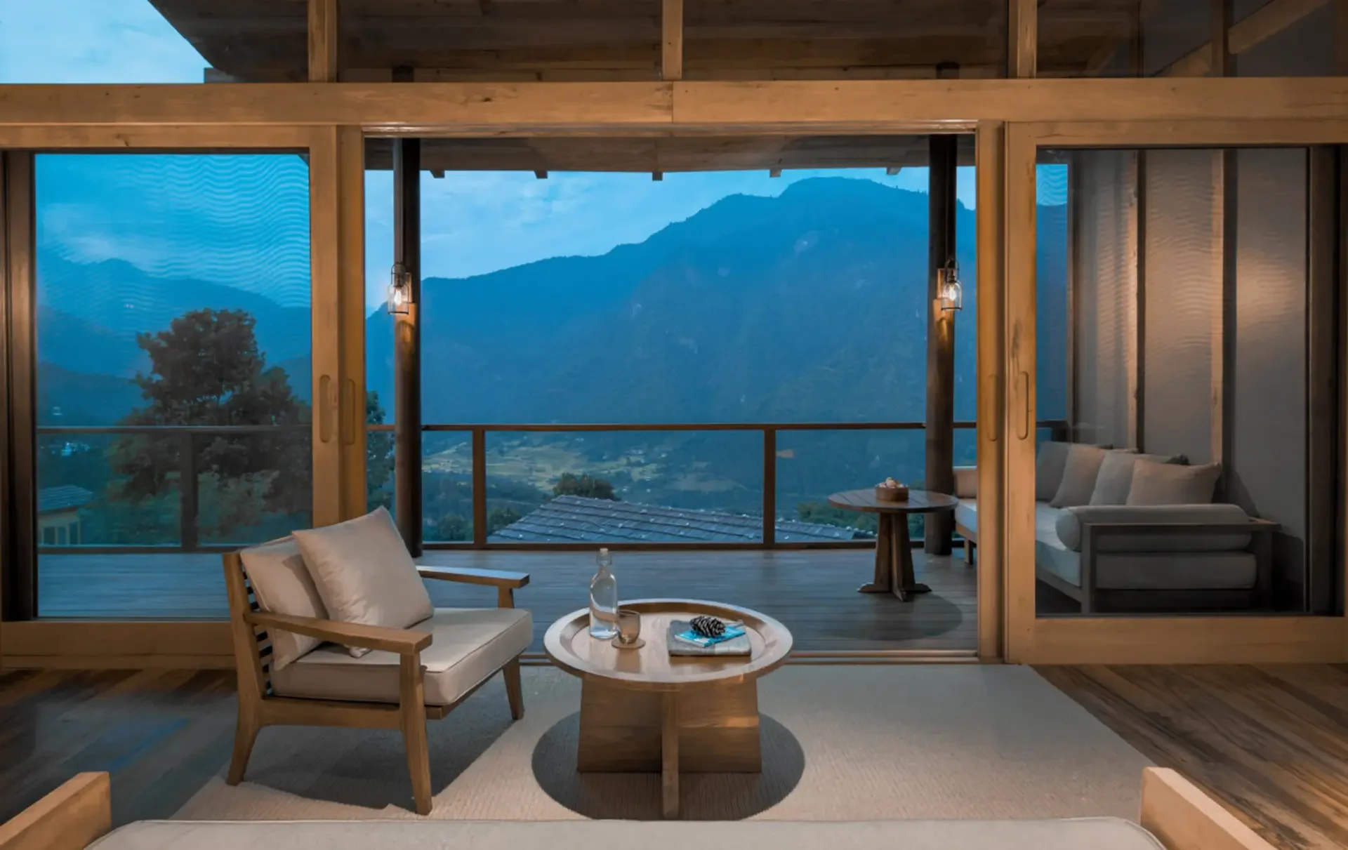 Living room with large windows overlooking mountains, featuring wooden furniture, a white sofa with cushions, a coffee table with water bottles and decorative items, and outdoor scenery.