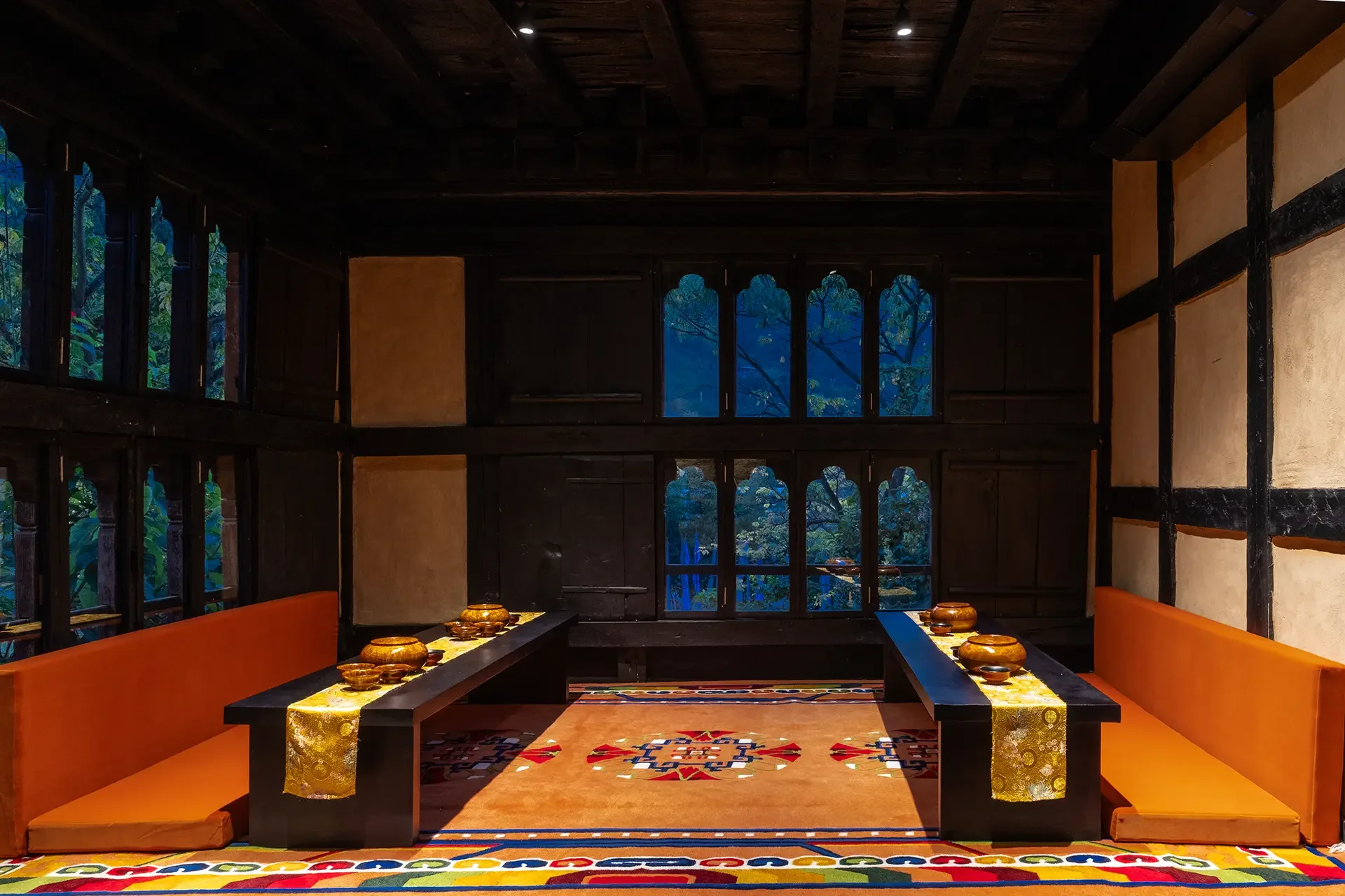 Interior of a traditional room with dark wood beams, large windows showing a view of trees outside, and orange seating along a black table with yellow tablecloth and decorative bowls.