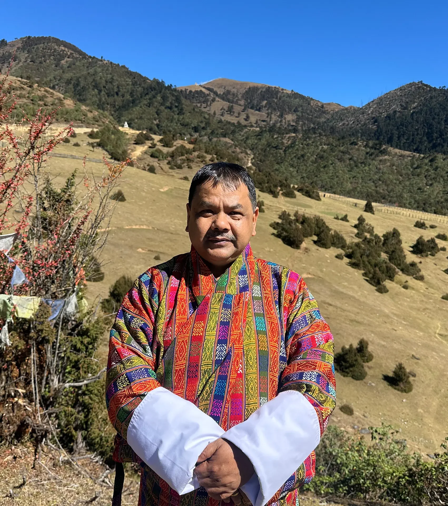 A man wearing a colorful traditional jacket stands outdoors in front of a mountainous landscape with blue sky, trees, and grassy hills.