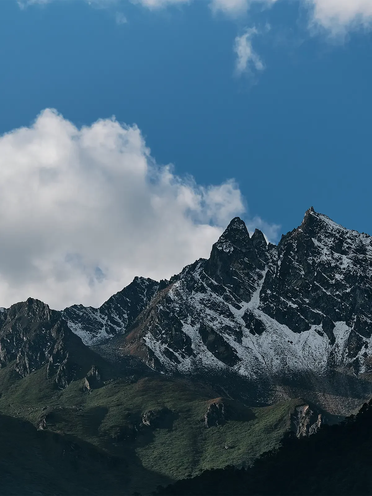 Snow-capped mountain peaks under a partly cloudy blue sky with green hills in the foreground.