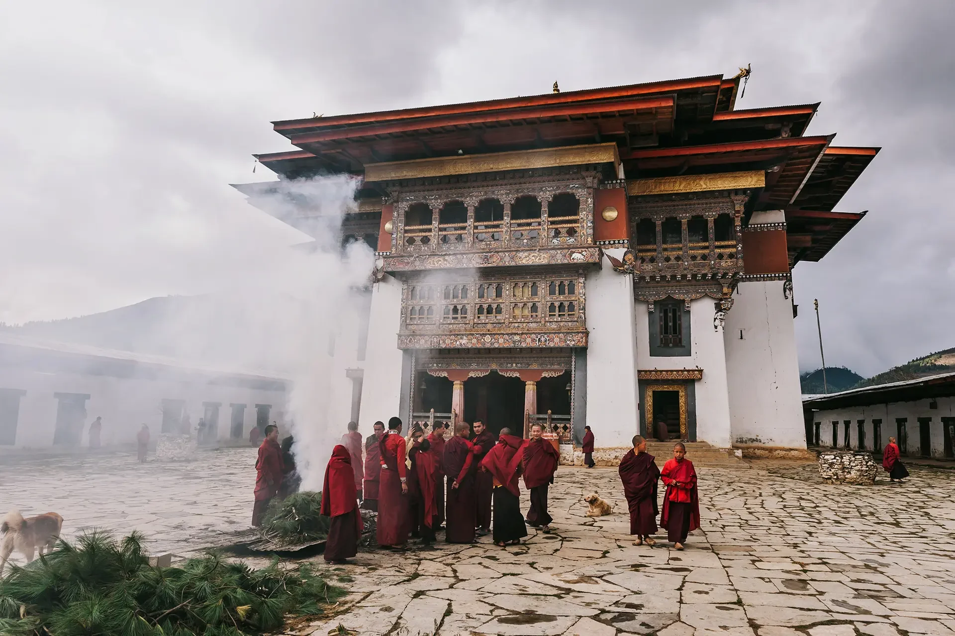 Buddhist monks in red robes gather outside a traditional Bhutanese temple with smoke rising from incense or ritual offerings, in a mountainous landscape under cloudy skies.