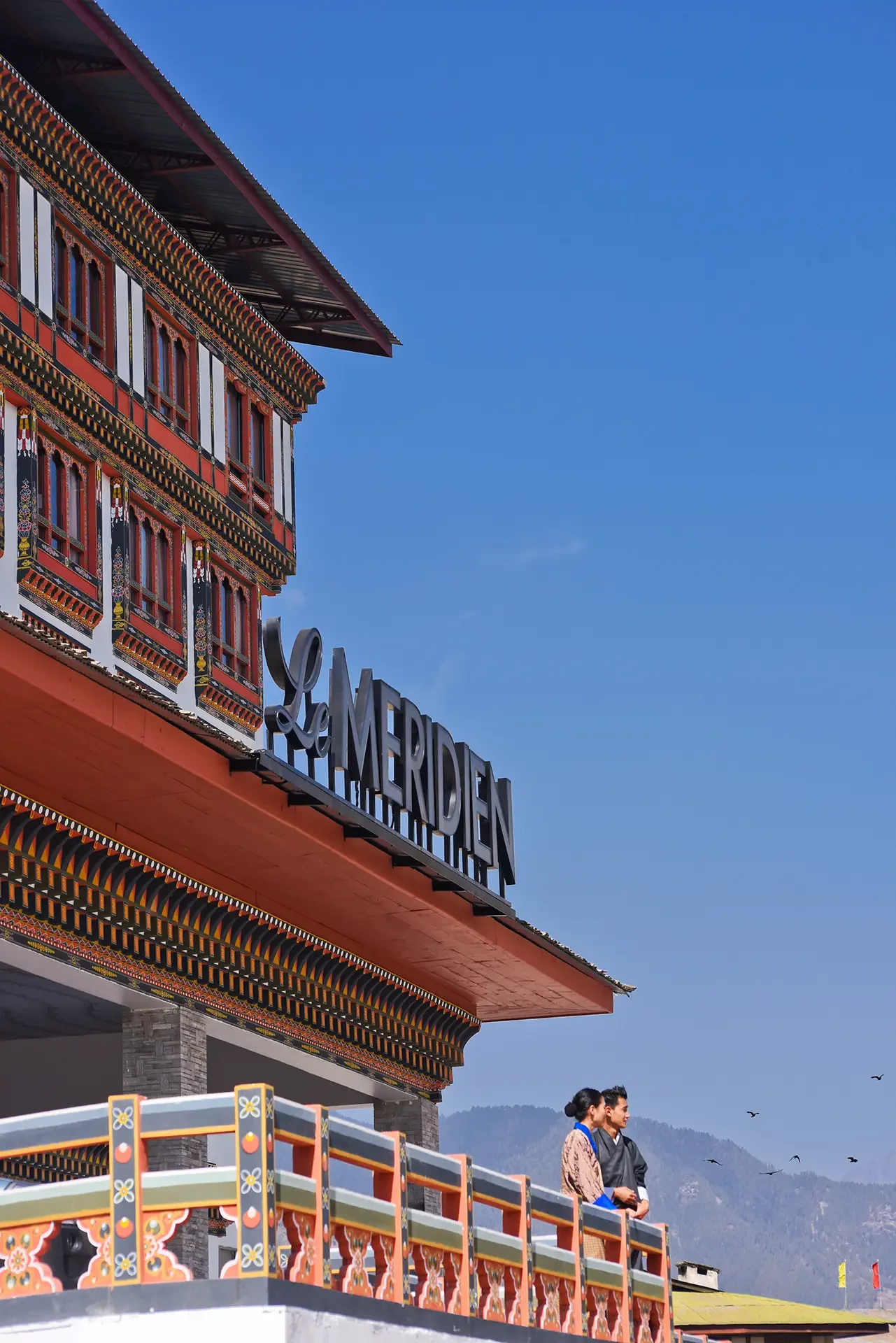 A colorful building with traditional Asian architectural details and the sign 'Meriden' in front of a mountain landscape, with two people dressed in traditional Asian clothing standing on a decorated balcony.