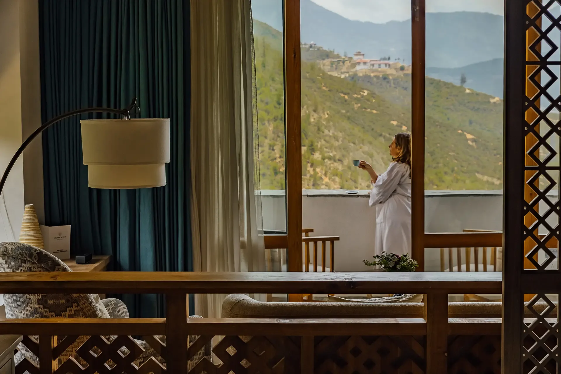 A woman in a white robe standing on a balcony, holding a cup and looking at the mountain landscape with houses and trees in the background.